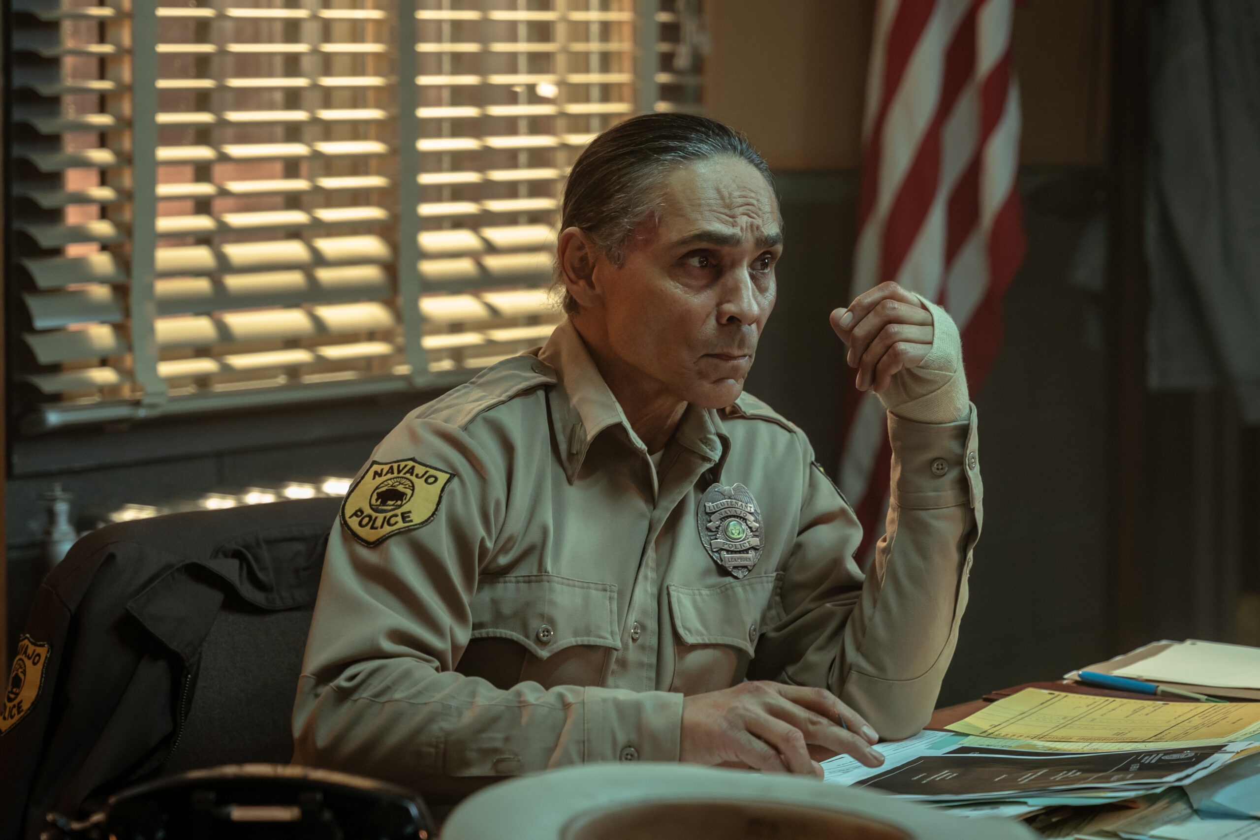 Dark Winds Season 2 Episode 5 - Zahn McClarnon | Tell-Tale TV Still from Dark Winds Season 2 Episode 5 of Zahn McClarnon as Joe Leaphorn sitting at a desk wearing a police uniform wihth one of his hand bandaged