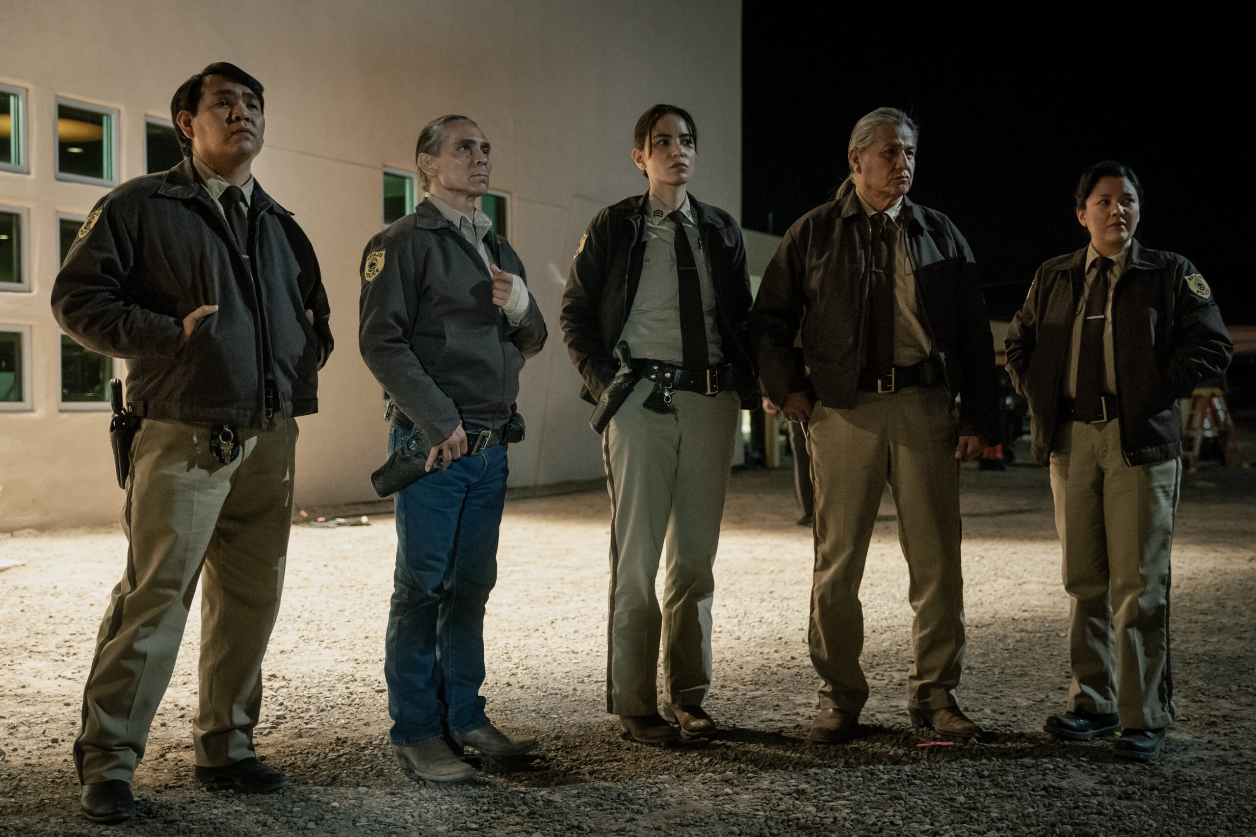 Dark Winds Season 2 Episode 5 - Zahn McClarnon, Jessica Matten, Andersen Kee, Wade Adakai,and Natalie Benally | Tell-Tale TV Still from Dark Winds Season 2 Episode 5 of Zahn McClarnon as Joe Leaphorn, Jessica Matten as Sgt. Bernadette Manuelito, Andersen Kee as Officer Harold Bigman, Wade Adakai as Officer Gary Felix and Natalie Benally as Natalie Bluehouse standing in a line outside the police station all wearing police uniforms