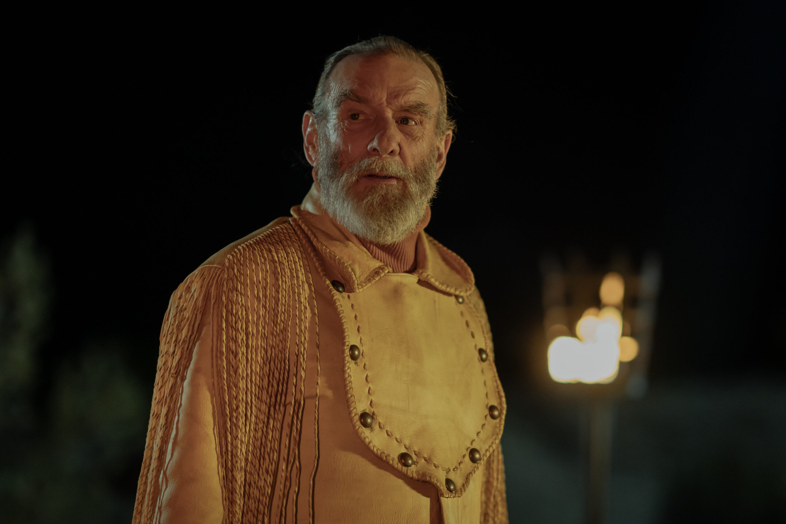 Dark Winds Season 2 Episode 5 - John Henry Diehl | Tell-Tale TV Still from Dark Winds Season 2 Episode 5 of John Henry Diehl as BJ Vines. Medium shot of Diehl wearing western style attire wth a blurred torch in the back ground