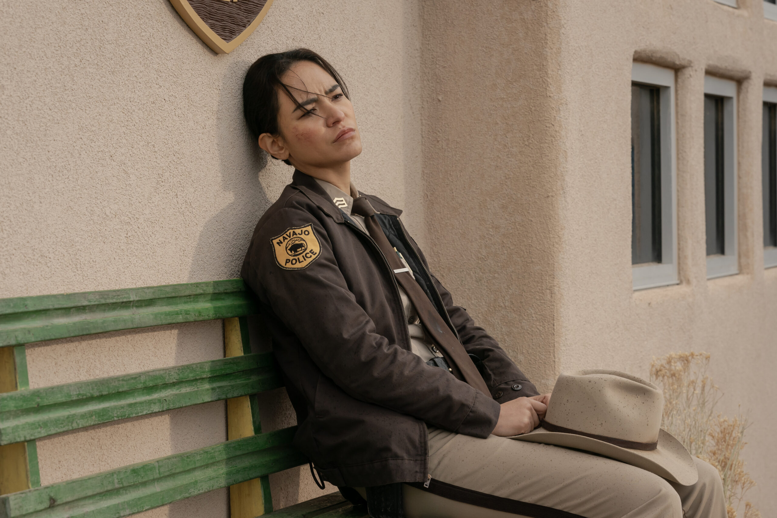 Dark Winds Season 2 Episode 5 - Jessica Matten | Tell-Tale TV Still from Dark Winds Season 2 Episode 5 of Jessica Matten as Sgt. Bernadette Manuelito sitting on a bench wearing her uniform