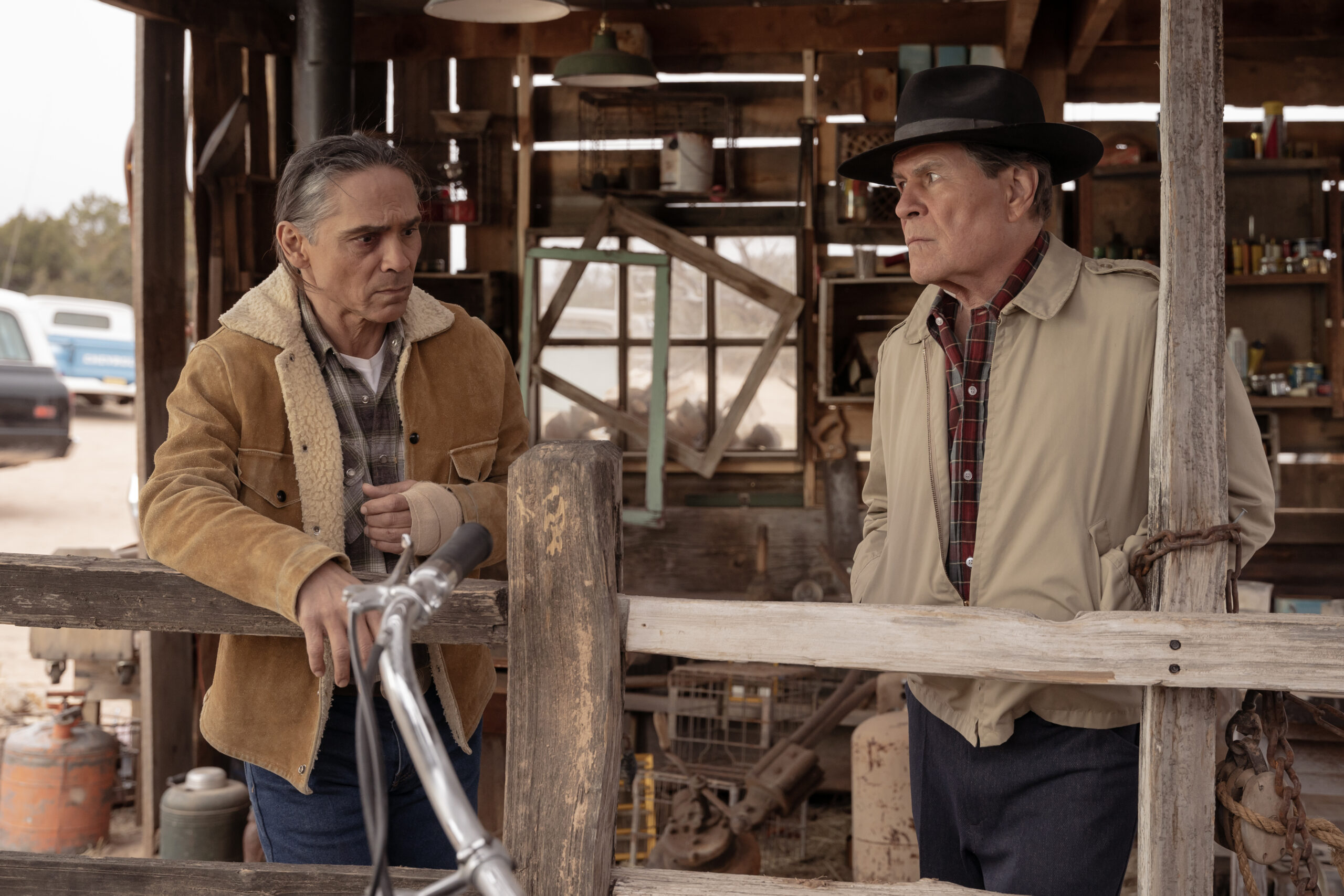 Dark Winds Season 2 Episode 6 - Zahn McClarnon and A. Martinez | Tell-Tale TV Still from Dark Winds Season 2 Episode 6 of Zahn McClarnon as Joe Leaphorn and A. Martinez as Gordo Sena standing in a wooden barn or shed behind a wooden fence