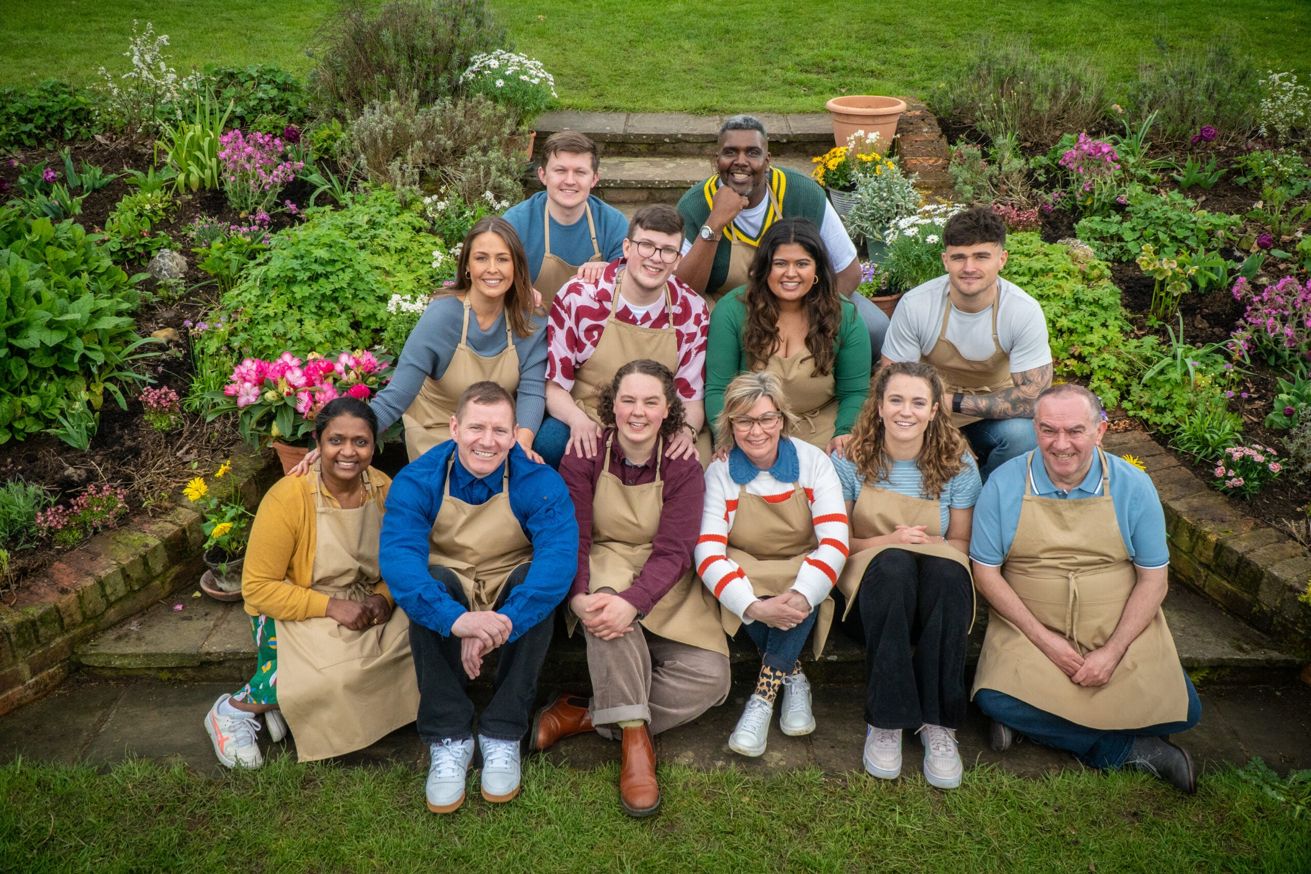 Production still from The Great British Bake Off Collection 11 of the season's contestants sitting in a group outside wearing tan aprons.