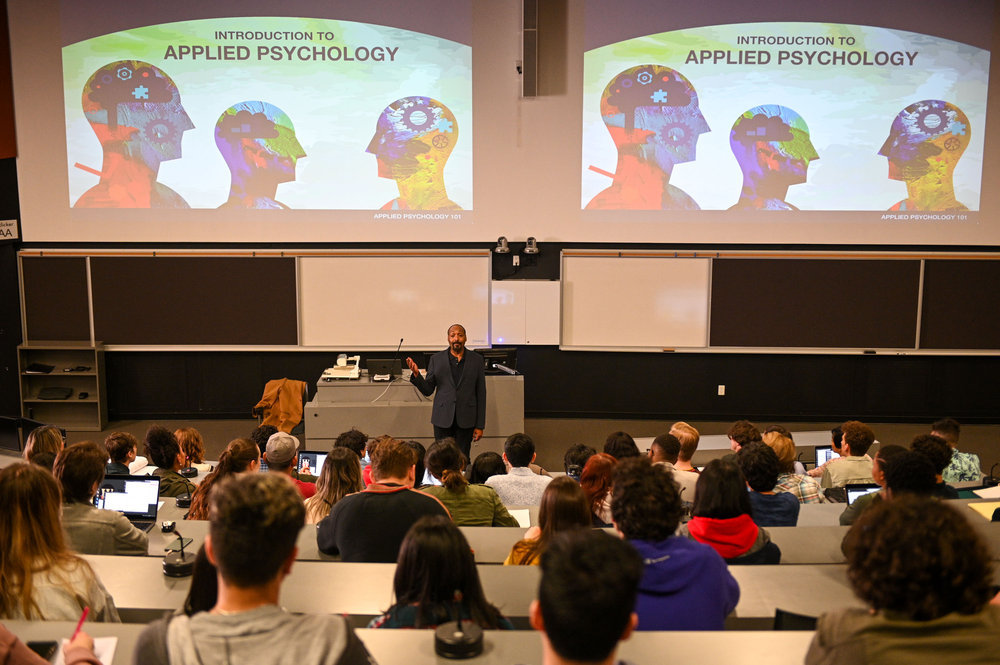 Still from The Irrational Season 1 Episode 1 of Martin as Alec Mercer standing in front of a large lecture class teaching a class with a slide on large screens that say Introduction to Applied Psychology