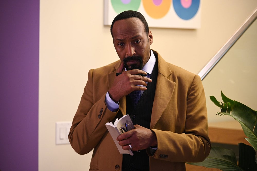 Still from The Irrational Season 1 Episode 1 of Jesse L. Martin as Alec Mercer. Medium shot of Martin in a tan trench coat holding a notebook and pen