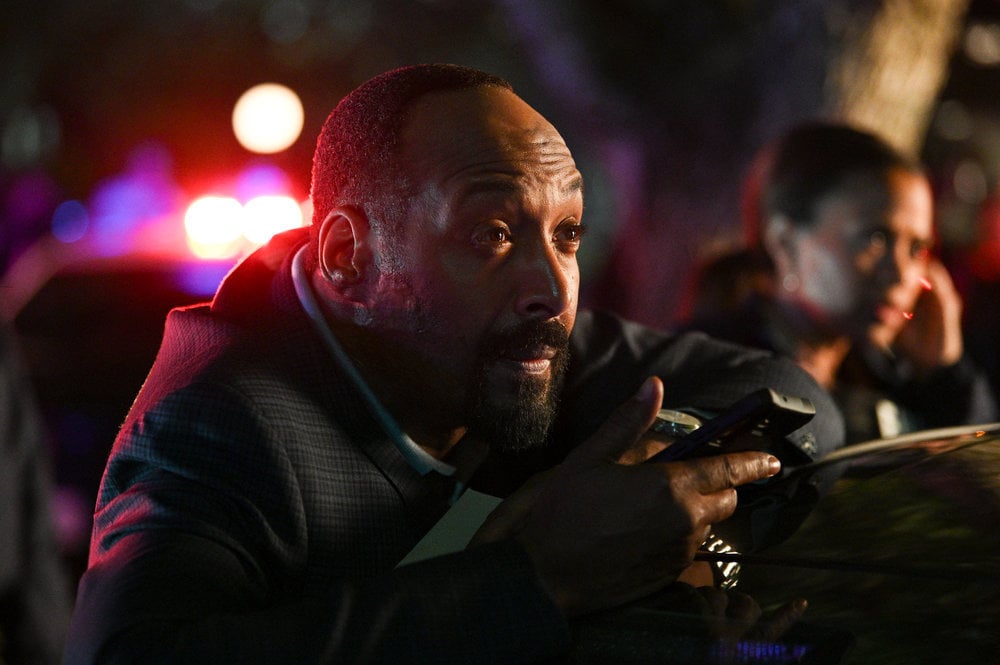 Still from The Irrational Season 1 Episode 1 of Jesse L. Martin as Alec Mercer, Maahra Hill as Marisa. Close shot of Martin holding a phone and standing behind a car with Hill blurred in the backgroud