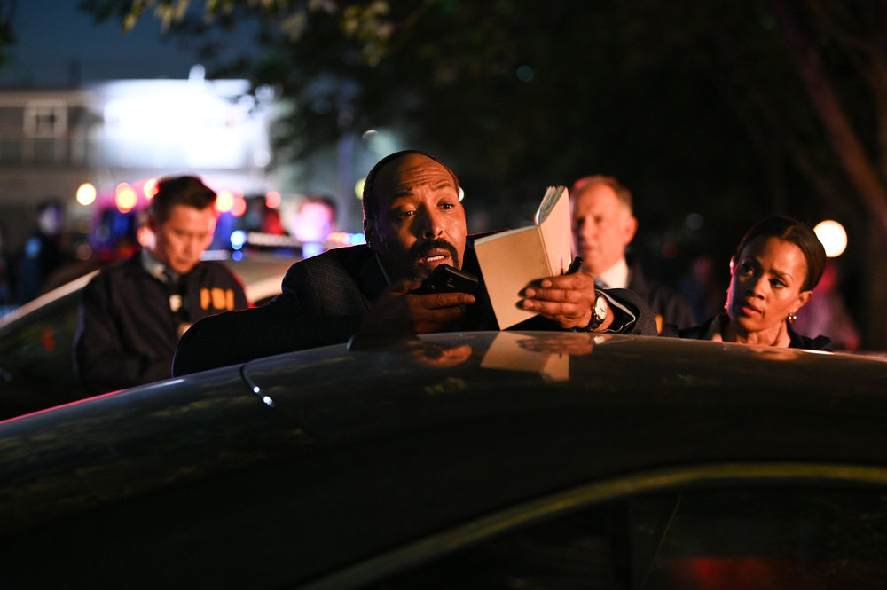 Still from The Irrational of Jesse L. Martin as Alec Mercer, Maahra Hill as Marisa at a night time outdoor crime scene wiith the pair standing behind a car and Martin holding a phone and notebook