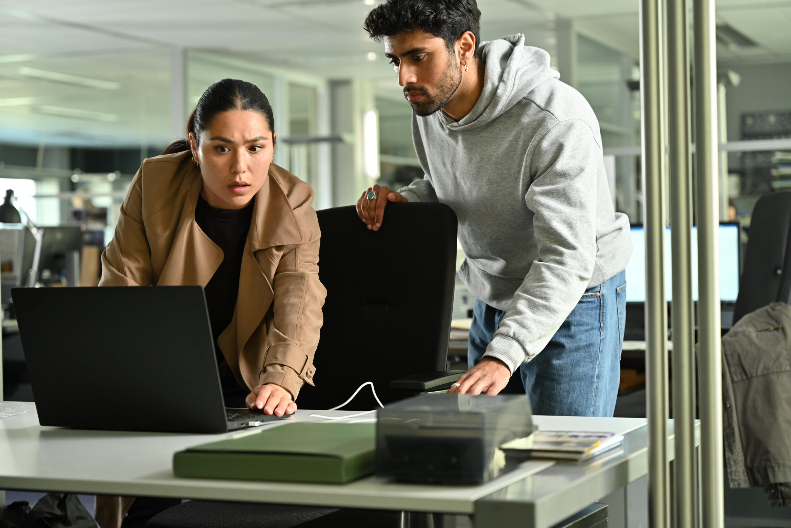 Van Der Valk Season 3 Episode 1 Part 2 - Django Chan-Reeves Li and Azan Ahmed | Tell-Tale TV Still from Van Der Valk SEason 3 Episode 1 Part 2. Photo of a man and a woman in an office looking at a lap top.