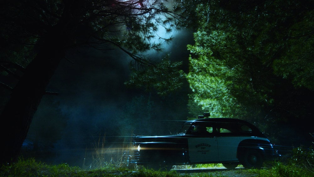 Still from Quantum Leap Season 2 Episode 3 of a Sheriff's Car driving on a dark road past some trees.