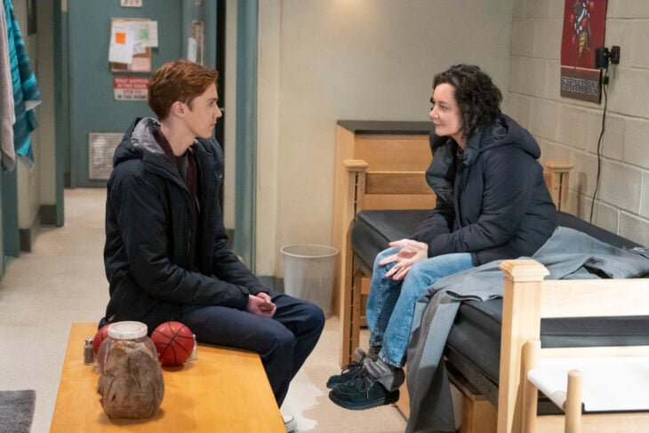 The Conners Season 6 Episode 4 - AMES MCNAMARA sits on a table in a college dorm while SARA GILBERT sits on the bed. They are facing each other talking.