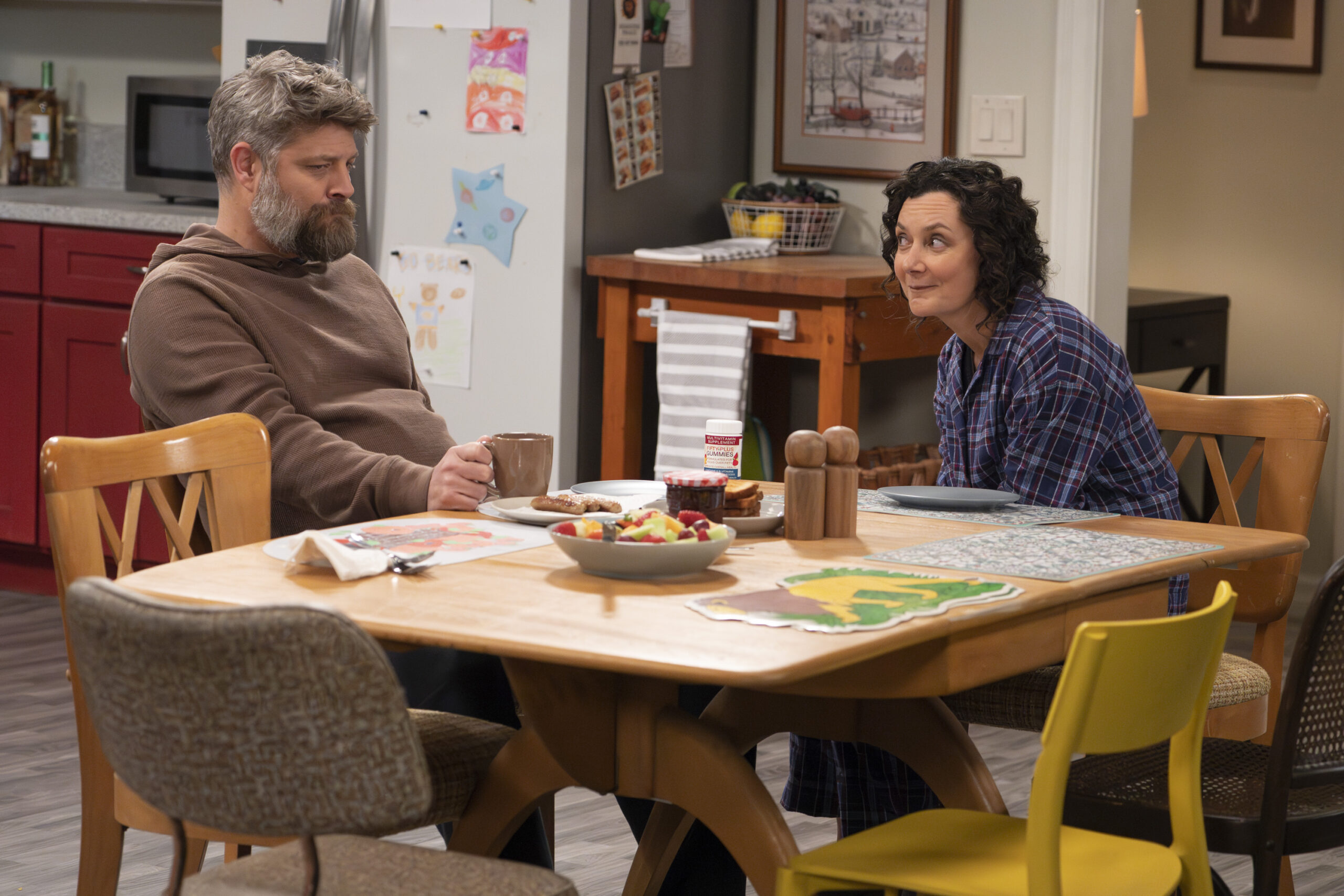The Conners Season 6 Episode 6 - JAY R. FERGUSON, SARA GILBERT sitting at their kitchen table. Darlene looks encouragingly at Ben as Ben sits and mopes with a coffee mug in hand.