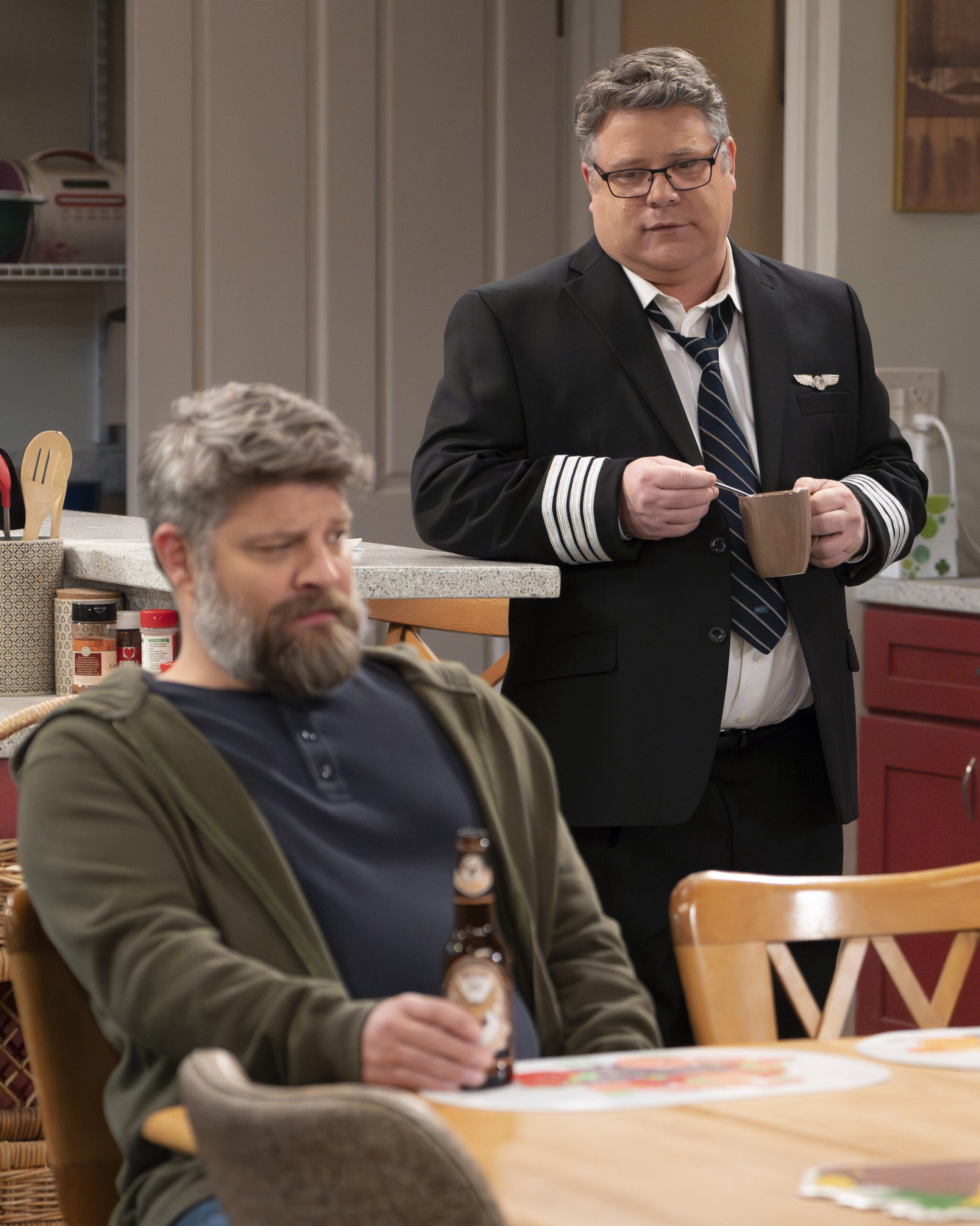 The Conners Season 6 Episode 6 - JAY R. FERGUSON, SEAN ASTIN in the kitchen of Darlene and Ben's home. Tyler stands at the counter behind Ben as he sits at the table.