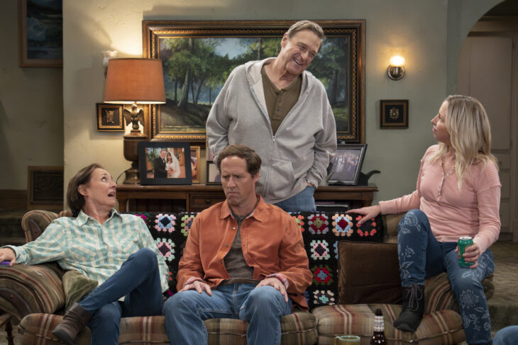 The Conners Season 6 Episode 5 - LAURIE METCALF, NAT FAXON, LECY GORANSON sit on the couch in the Conner home while JOHN GOODMAN stands behind the couch smiling at LECY.