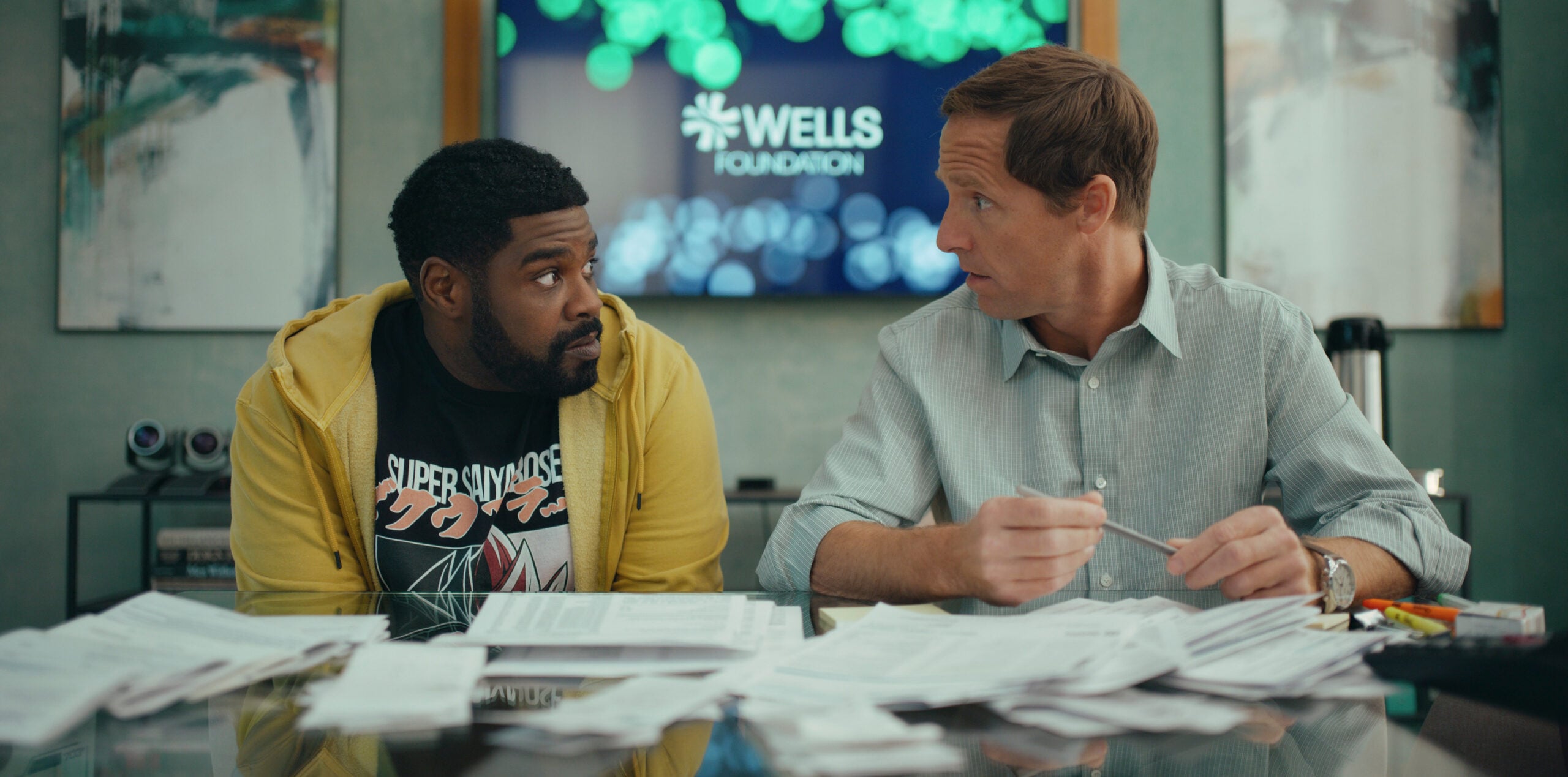 Still from Loot Season 2 Episode 2 of Ron Funches wearing a black graphic t-shirt and yellow hoodie and Nat Faxon wearing a light blue shirt. They are sitting at a desk with lots of papers in front of them from left to right.