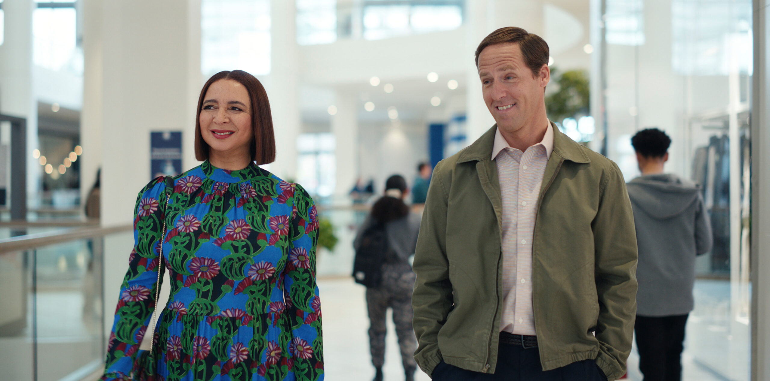 Loot Season 2 Episode 5 - Maya Rudolph and Nat Faxon | Tell-Tale TV Still from Loot Season 2 Episode 5 of Maya Rudolph wearing blue, green and black dress with purple flowers and Nat Faxon wearing a tan jacket over a button up shirt. They are standing next to each other form left to right.