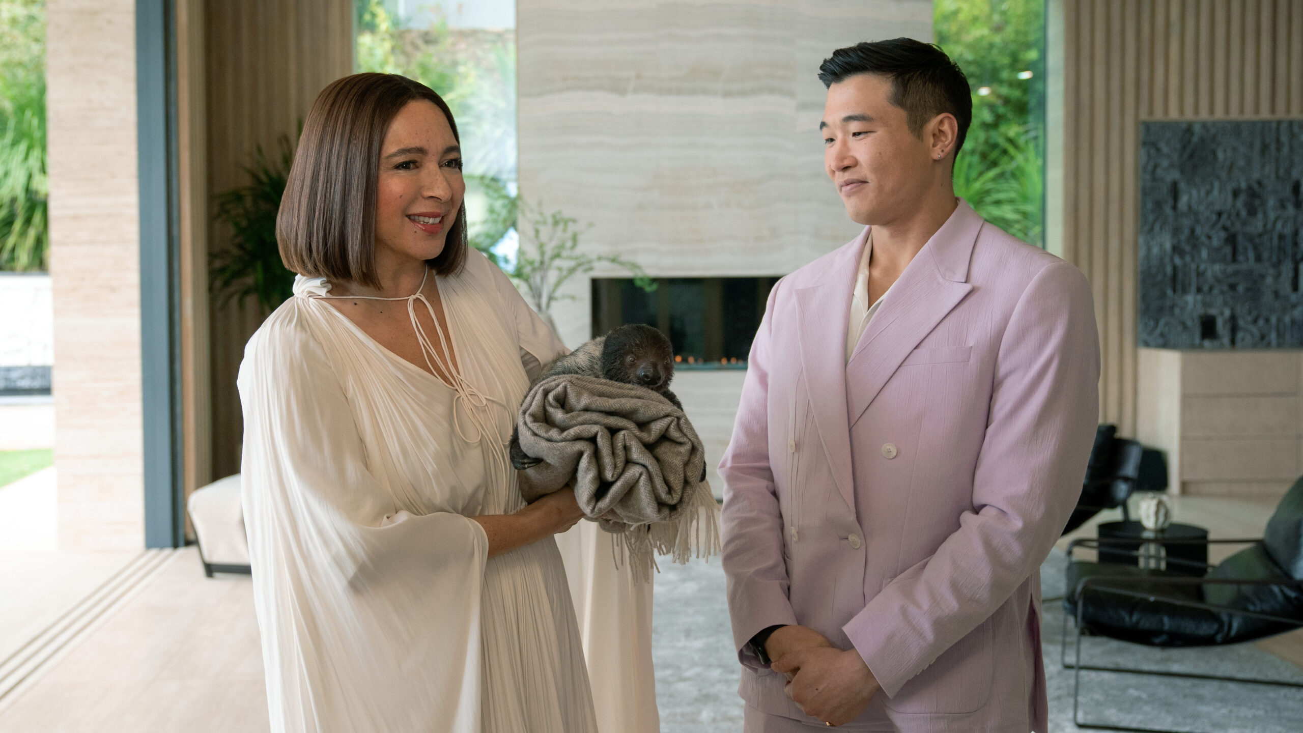 Still from Loot Season 2 Episode 1 of Maya Rudolph wearing a loose white cotton dress and holding a bundle of clothe and Joel Kim Booster wearing a pale pink jacket pictured from left to right.