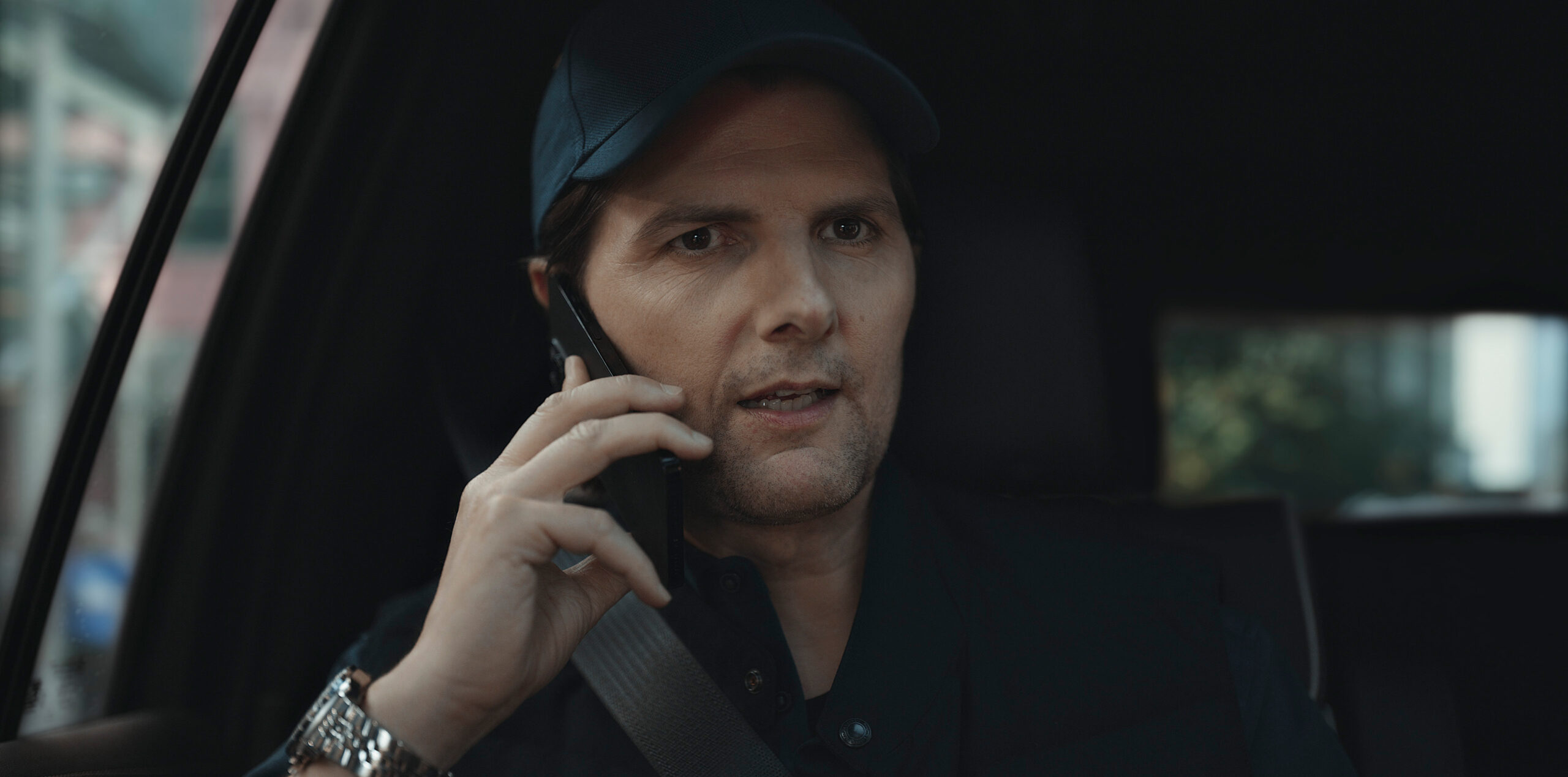 Still from Loot Season 2 Episode 1 of Adam Scott in a close up wearing black sitting in a car talking on a cell phone.