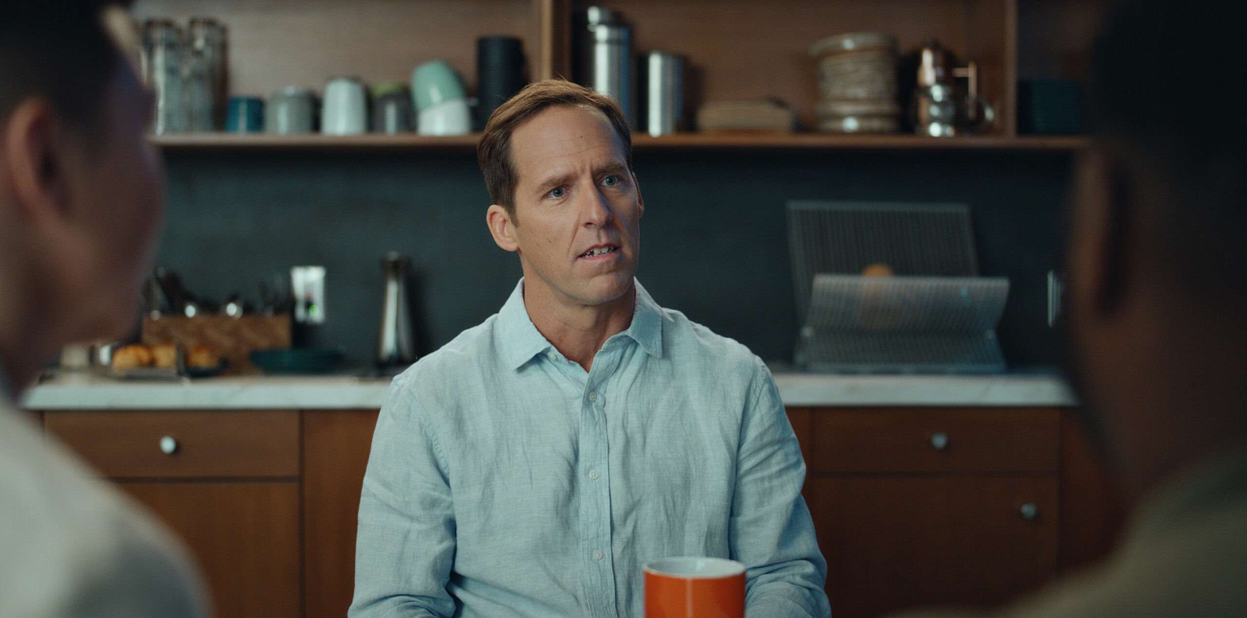 Still from Loot Season 2 Episode 1 of Nat Faxon in a medium shot sitting in a kitchen wearing a light blue shirt.