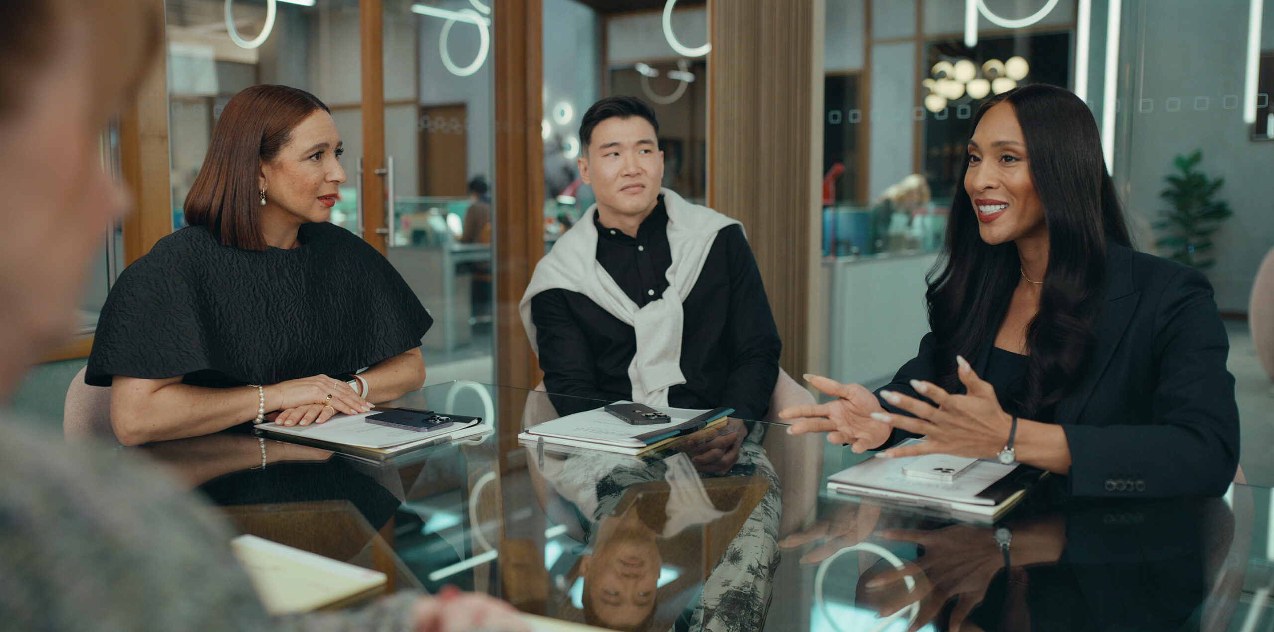 Still from Loot Season 2 Episode 2 of Maya Rudolph, Joel Kim Booster and Michaela Jaé Rodriguez sitting at a conference table from left to right.