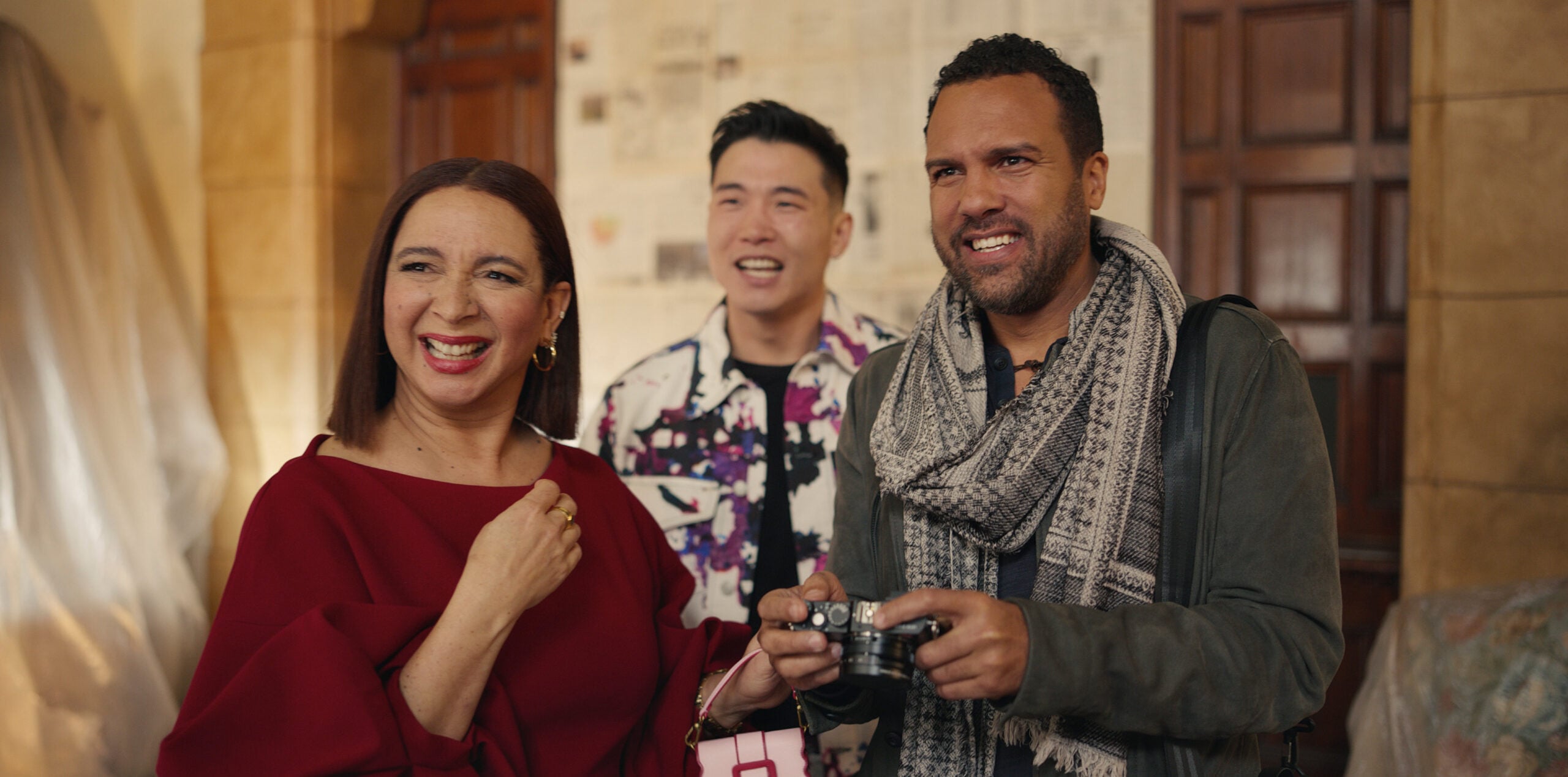 Still from Loot Season 2 Episode 2 of Maya Rudolph in a maroon top smiling, Joel Kim Booster in a black t-shirt with a patterned white, black, purple, and pink jacket over it smiling, and O-T Fagbenle wearing a gray jacket and scarf and holding a camera. They are pictured from left to right.
