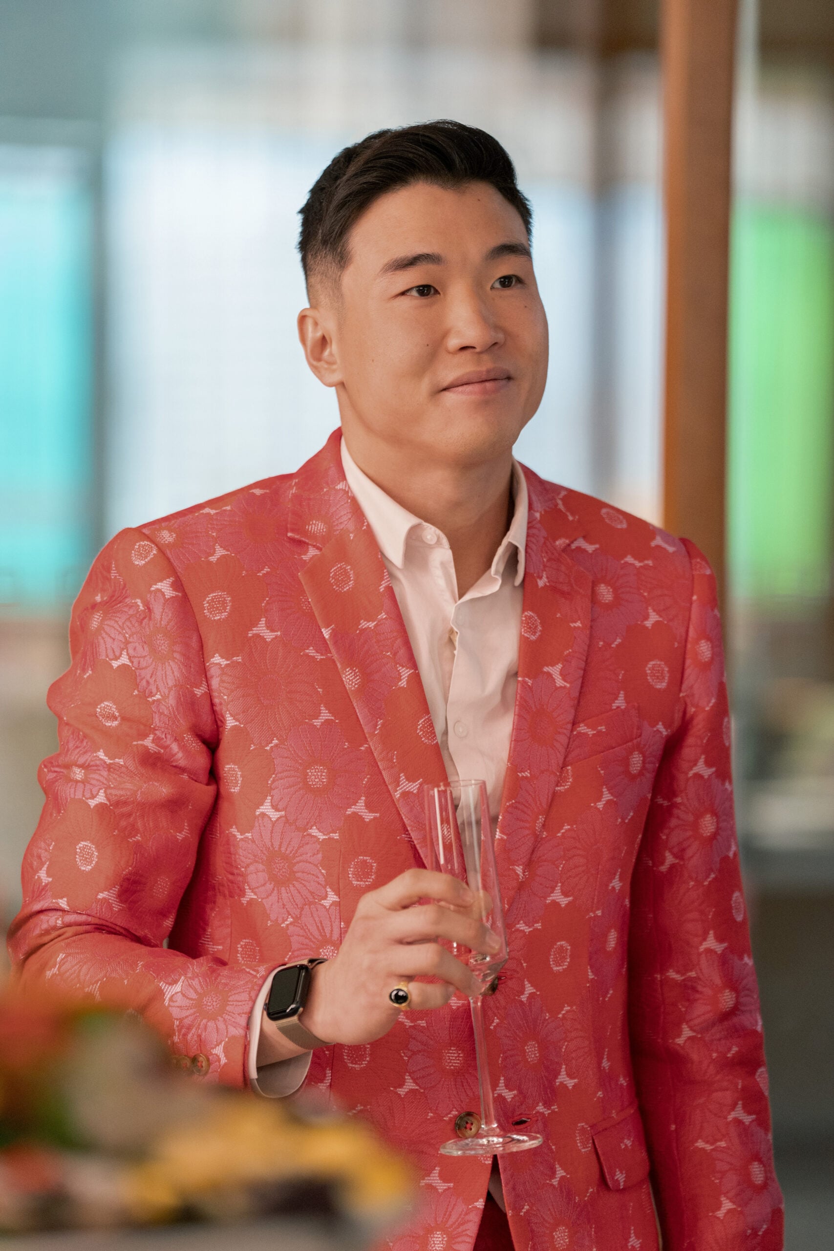 Still from Loot Season 2 Episode 2 of Joel Kim Booster wearing an orange shirt with a flowered pattern on the jacket and a light pink shirt under the jacket. He is holding a champagne glass.