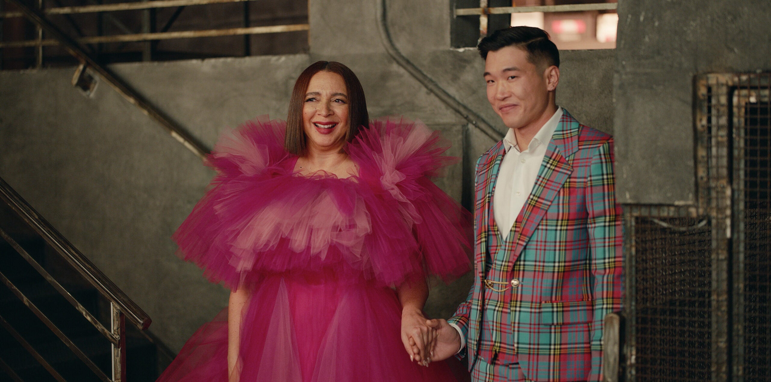 Still from Loot Season 2 Episode 6 of Maya Rudolph wearing a ver frilly pink dress and Joel Kim Booster wearing a blue, red, green and yellow plaid suit. They are holding hands and standing left to right.