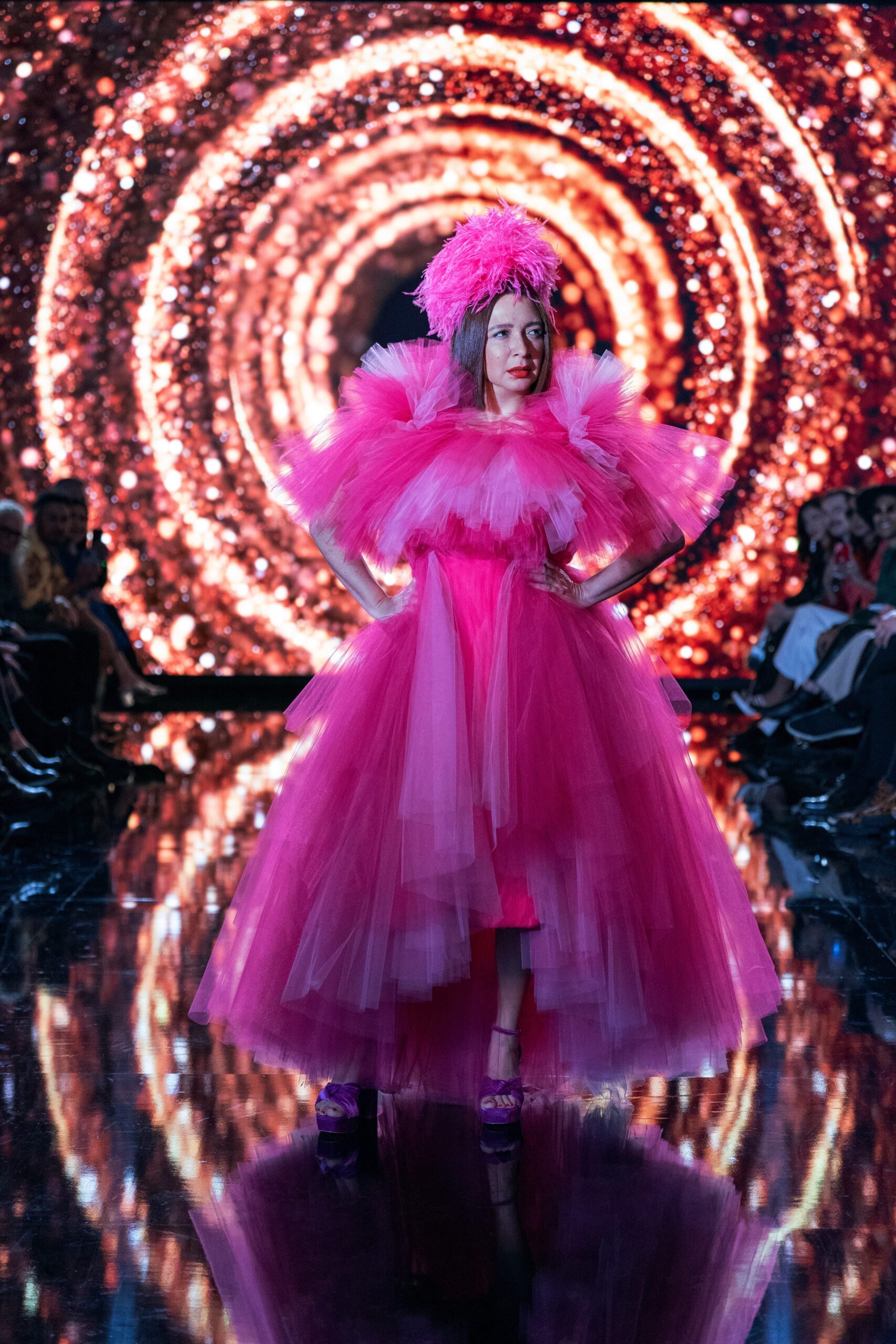 Still from Loot Season 2 Episode 6 of Maya Rudolph standing on a stage with circular lights in the back. She is wearing a very frilly pink dress and feathered pink hat.
