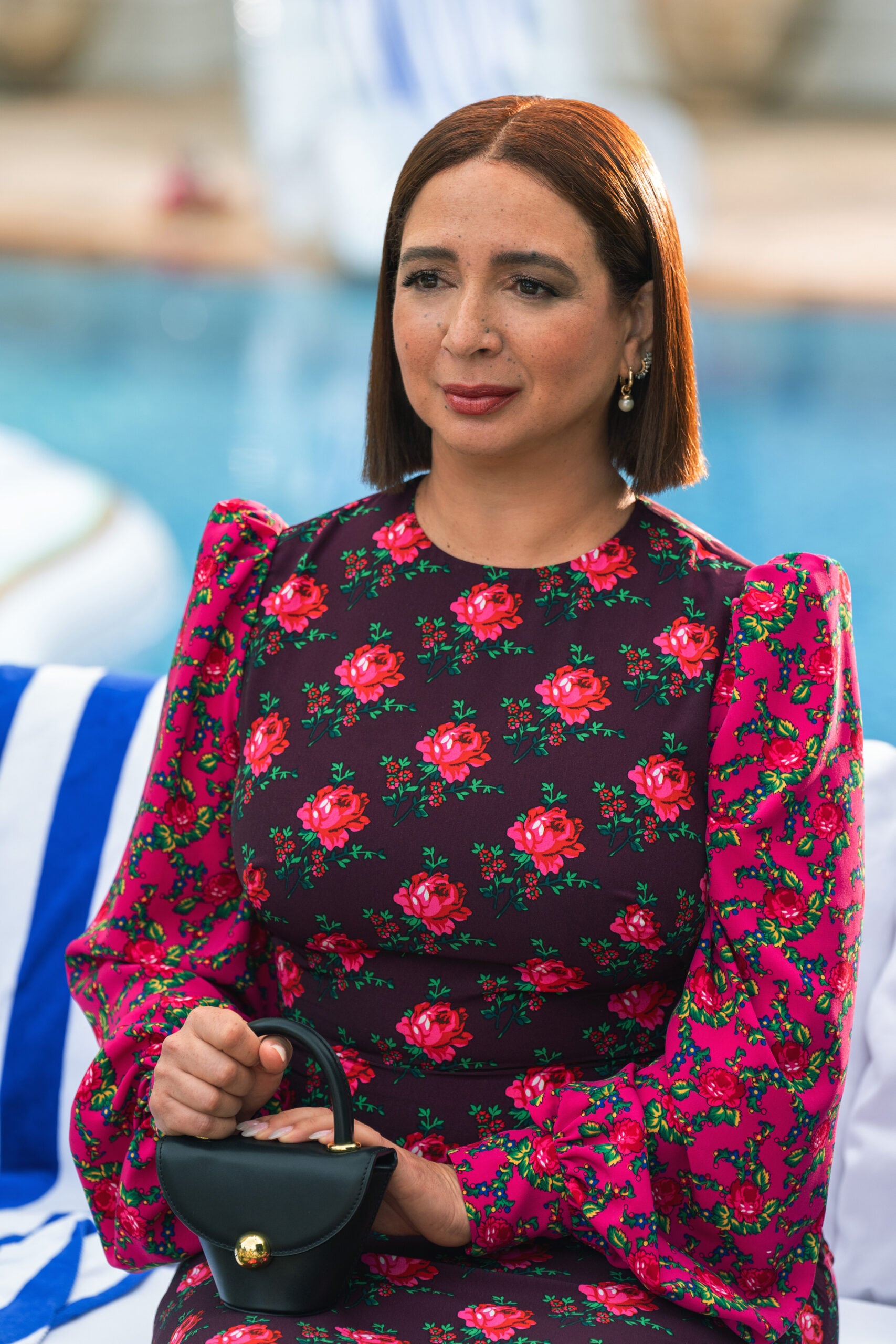 Still from Loot Season 2 Episode 8 of Maya Rudolph sitting in front of a pool wearing a long sleeve shirt with flowers on it and holding a small purse.