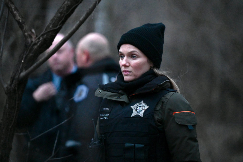 Chicago P.D. Season 11 Episode 7, Tracy Spiridakos as Hailey Upton stands wearing a police vest and beanie in a wooded area in the early hours.