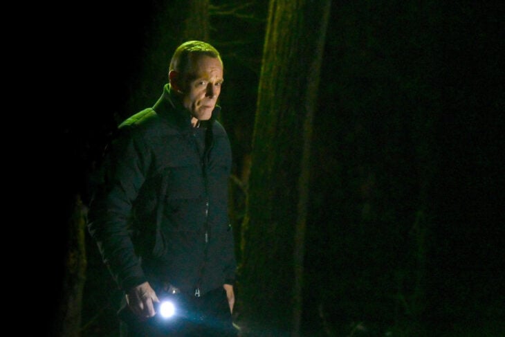 Chicago P.D. Season 11 Episode 7, Jason Beghe as Hank Voight walks through a wooded area at night with a flashlight looking concerned.