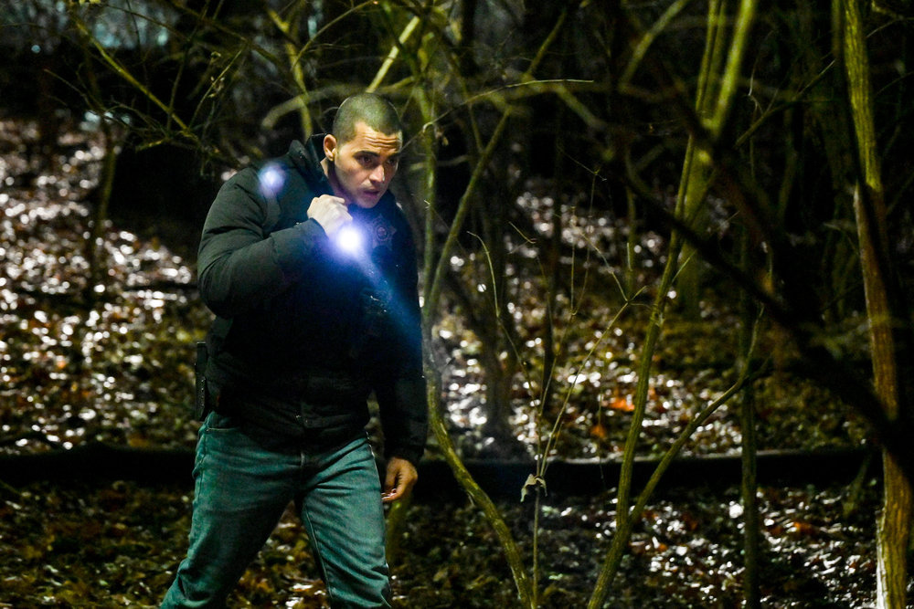 Chicago P.D. Season 11 Episode 7, Benjamin Levy Aguilar as Dante Torres walks through a wooded area at night wearing a police vest and shining a flashlight.