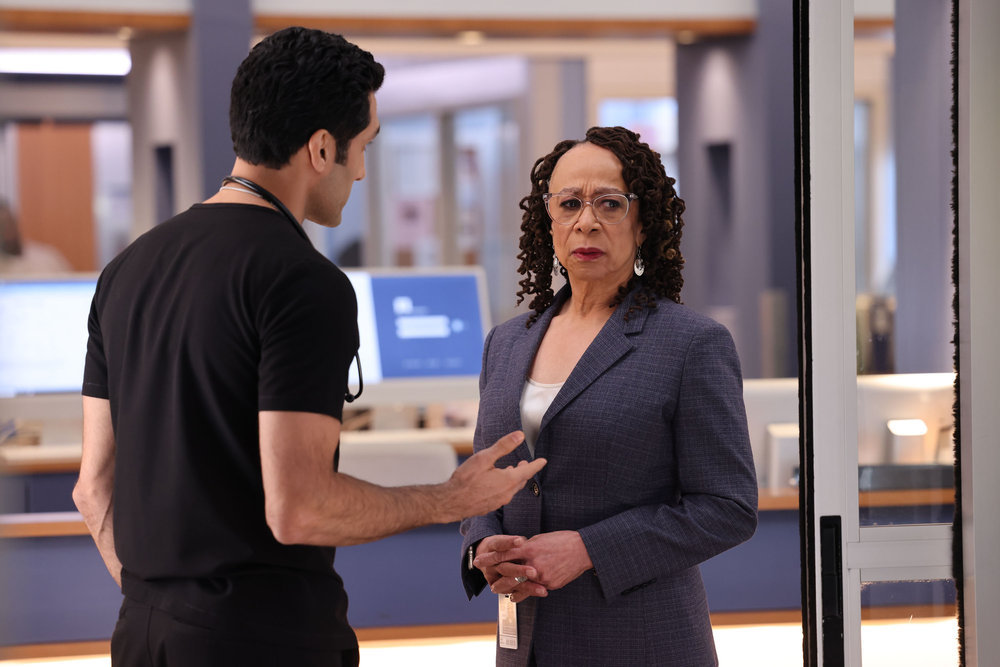 Chicago Med Season 9 Episode 7, Dominic Raines as Dr. Crockett Marcel stands with his back to the camera, and his right arm extended, addressing S. Epatha Merkerson as Sharon Goodwin who is looking concerned toward something out of frame.