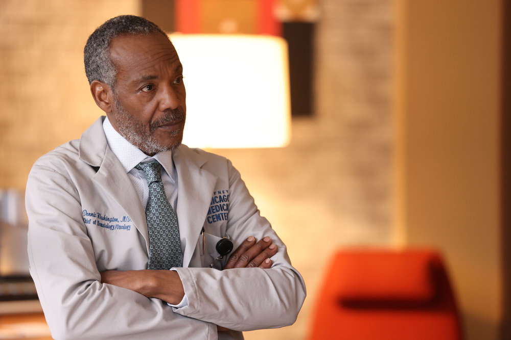 Chicago Med Season 9 Episode 7, John Earl Jenks as Dr. Dennis Washington stands with his arms crossed and an unamused look on his face.