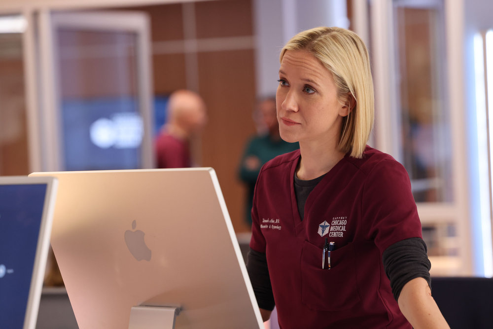Chicago Med Season 9 Episode 7. Jessy Schram as Dr. Hannah Asher stands at the computer looking to someone out of frame.