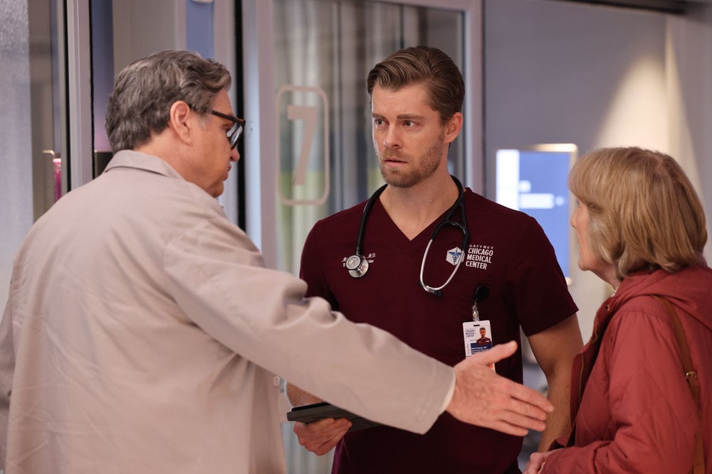 Chicago Med Season 9 Episode 7, Oliver Platt as Dr. Daniel Charles stands with his back mostly to the camera, extending his hand to an elderly female patient while Luke Mitchell as Dr. Mitch Ripley looks at him with concern on his face.