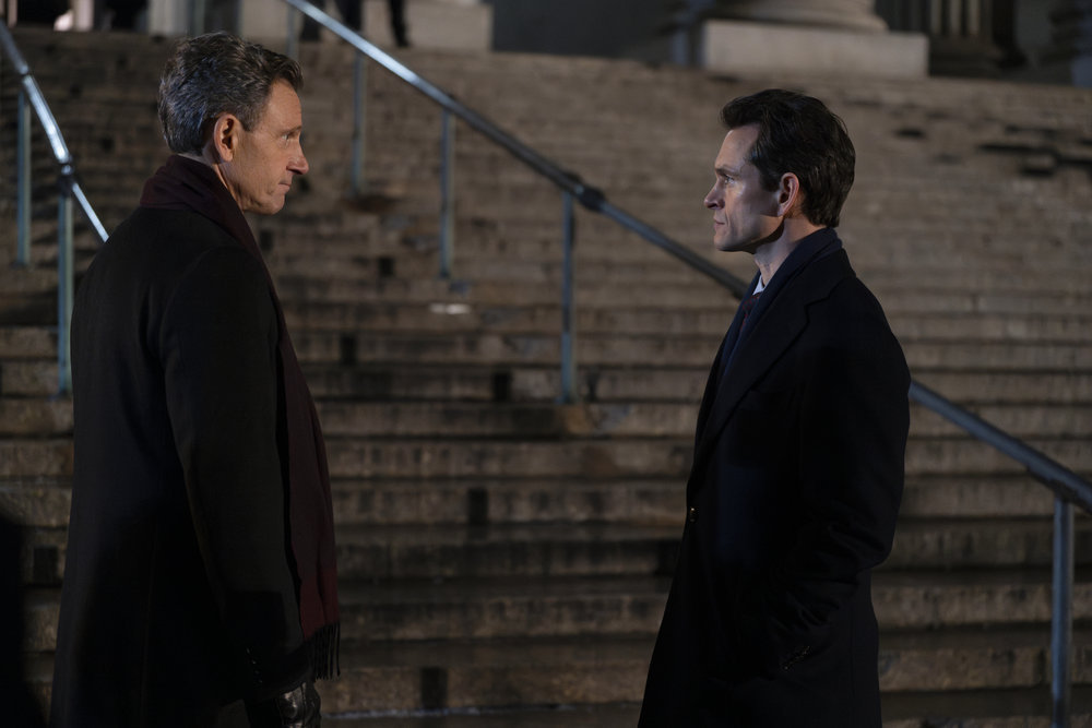 Law & Order - Season 23 | Tell-Tale TV Law & Order Season 23 Episode 8 - Tony Goldwyn and Hugh Dancy stand with their side profile facing the camera as they converse with each other at the bottom of the courthouse steps at night.