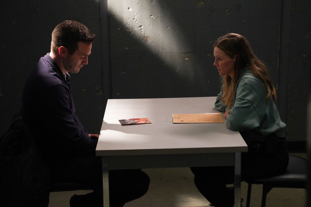 Chicago P.D. - Season 11 Episode 8 Andy Favreau as Trent Howell sits on one side of an interrogation table while Tracy Spiridakos as Hailey Upton sits across from him looking at him as he looks at a photograph on the table in front of him.