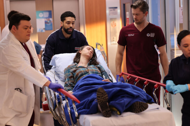 Chicago Med Season 9 Episode 9 - Conor Perkins as Dr. Zach Hudgens, Henderson Wade as Dr. Loren Johnson, and Luke Mitchell as Dr. Mitch Ripley escort a young white female patient on a gurney down the hall.