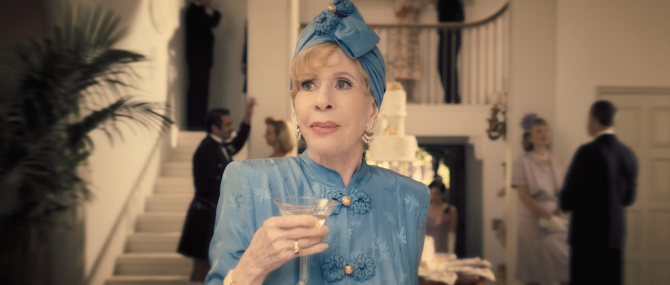 Norma wears a light blue silk dress with a matching blue silk head wrap. She holds a cocktail glass while standing among the crowd of a wedding party in Palm Royale Season 1 Episode 4, "Maxine Rolls the Dice."