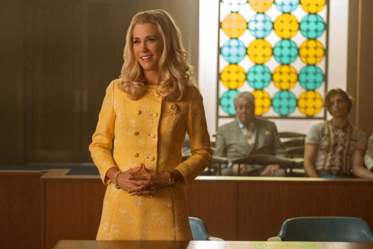 Maxine wears a yellow suit dress while standing in a courtroom. She faces away from the judge with her hands clasped in front of her while smiling in Palm Royale Season 1 Episode 4, "Maxine Rolls the Dice."