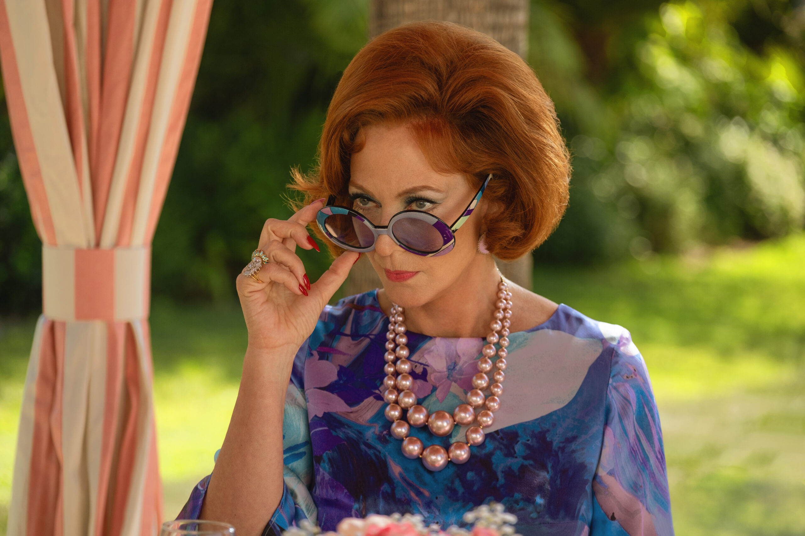 Evelyn pulls her sunglasses down while sitting under a canopy near a pool. She wears a bright blue and purple patterned dress on Palm Royale Season 1 Episode 1, "Pilot."