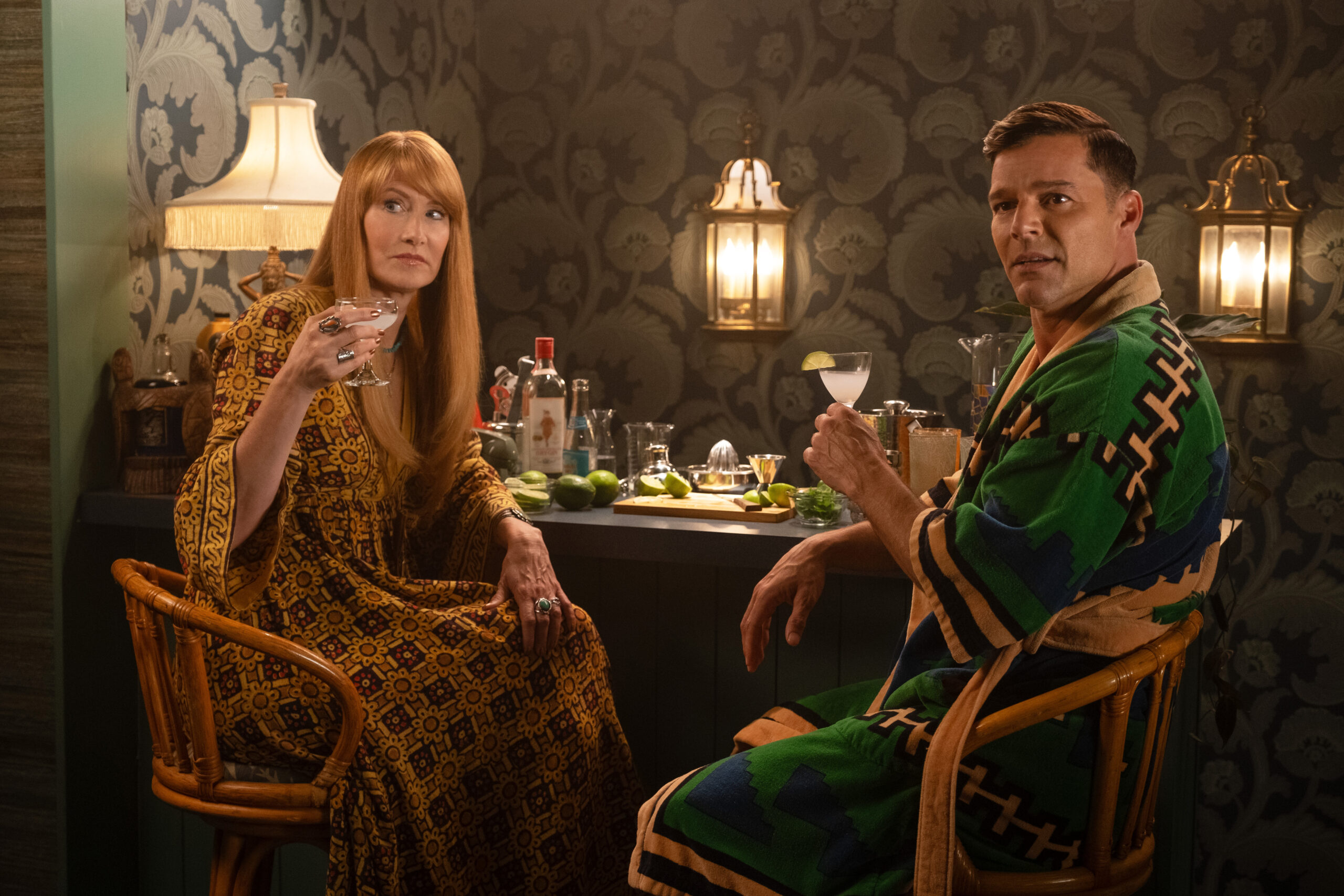 Linda wears a mustard yellow caftan dress while sitting at a bar next to Robert, who wears a green silk robe. Both have drinks in hand on Palm Royale Season 1 Episode 3, "Maxine's Like a Dellacorte." 