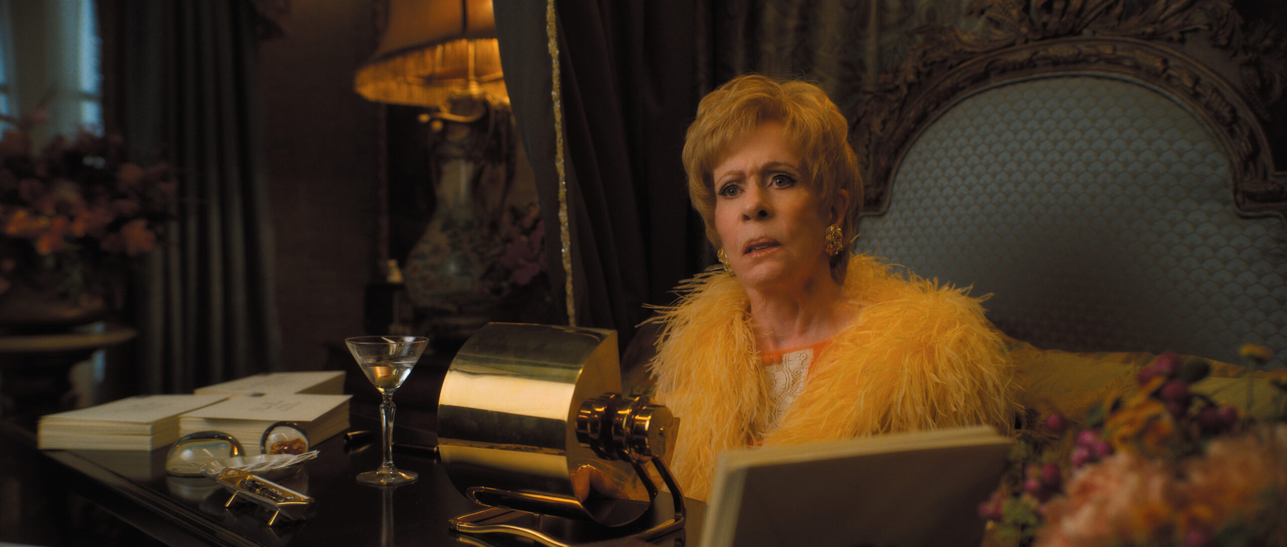 Norma wears a yellow feathered silk robe while sitting on her bed in her bedroom, a golden Rolodex before her, on Palm Royale Season 1 Episode 3, "Maxine's Like a Dellacorte." 