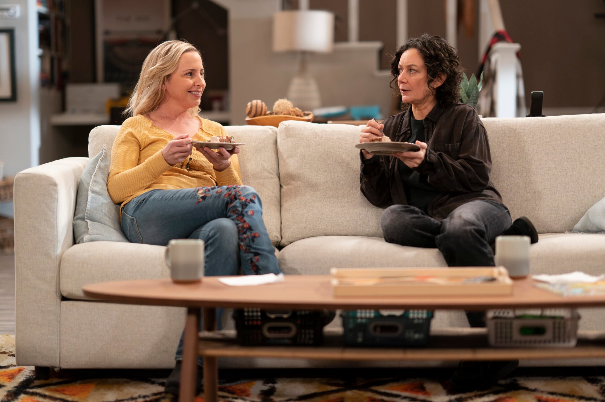 The Conners Season 6 Episode 7: LECY GORANSON and SARA GILBERT sit on their couch eating and chatting.