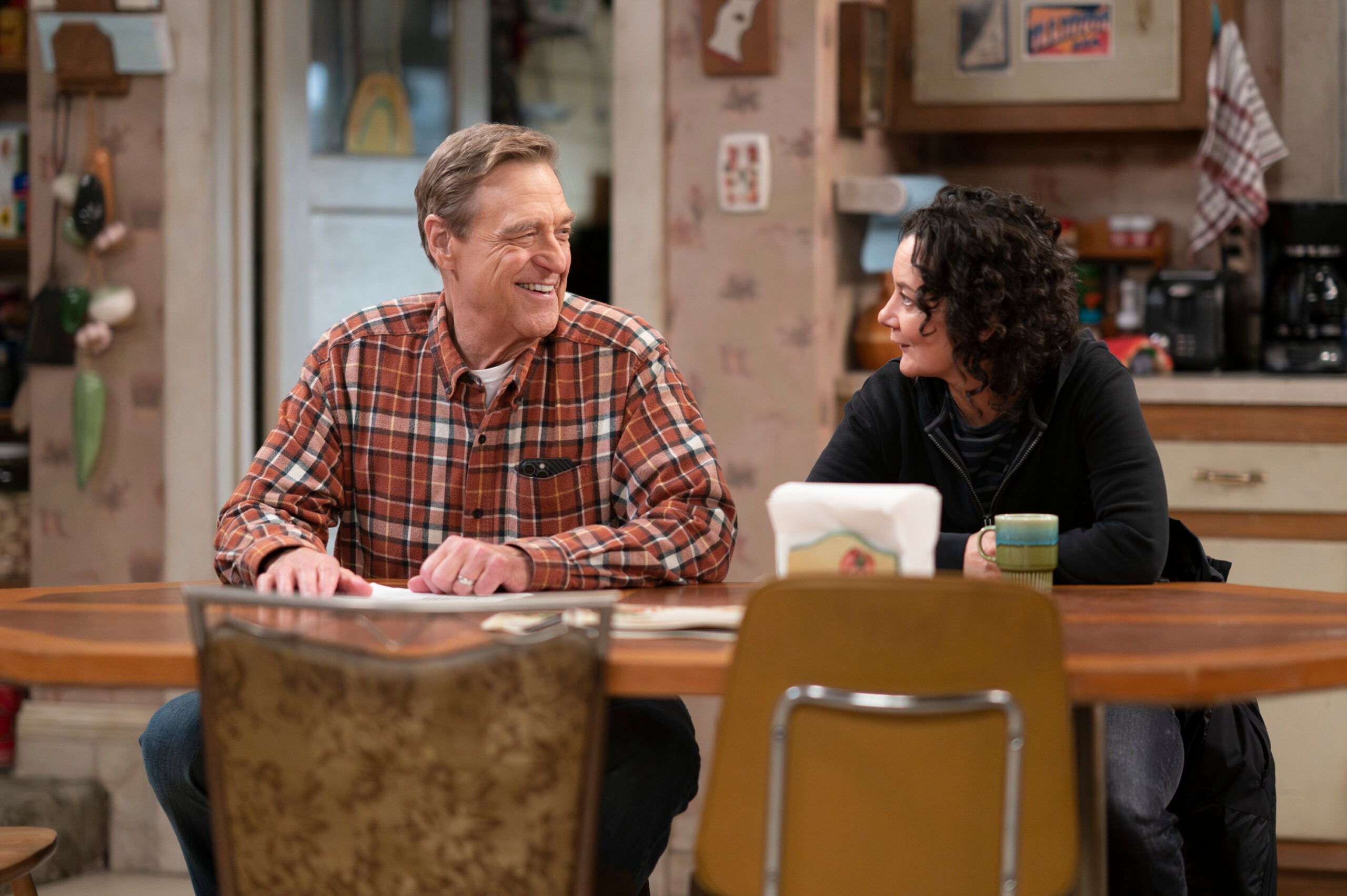 The Conners Season 6 Episode 7: JOHN GOODMAN, SARA GILBERT sit at the Conner kitchen table, smiling at each other. 