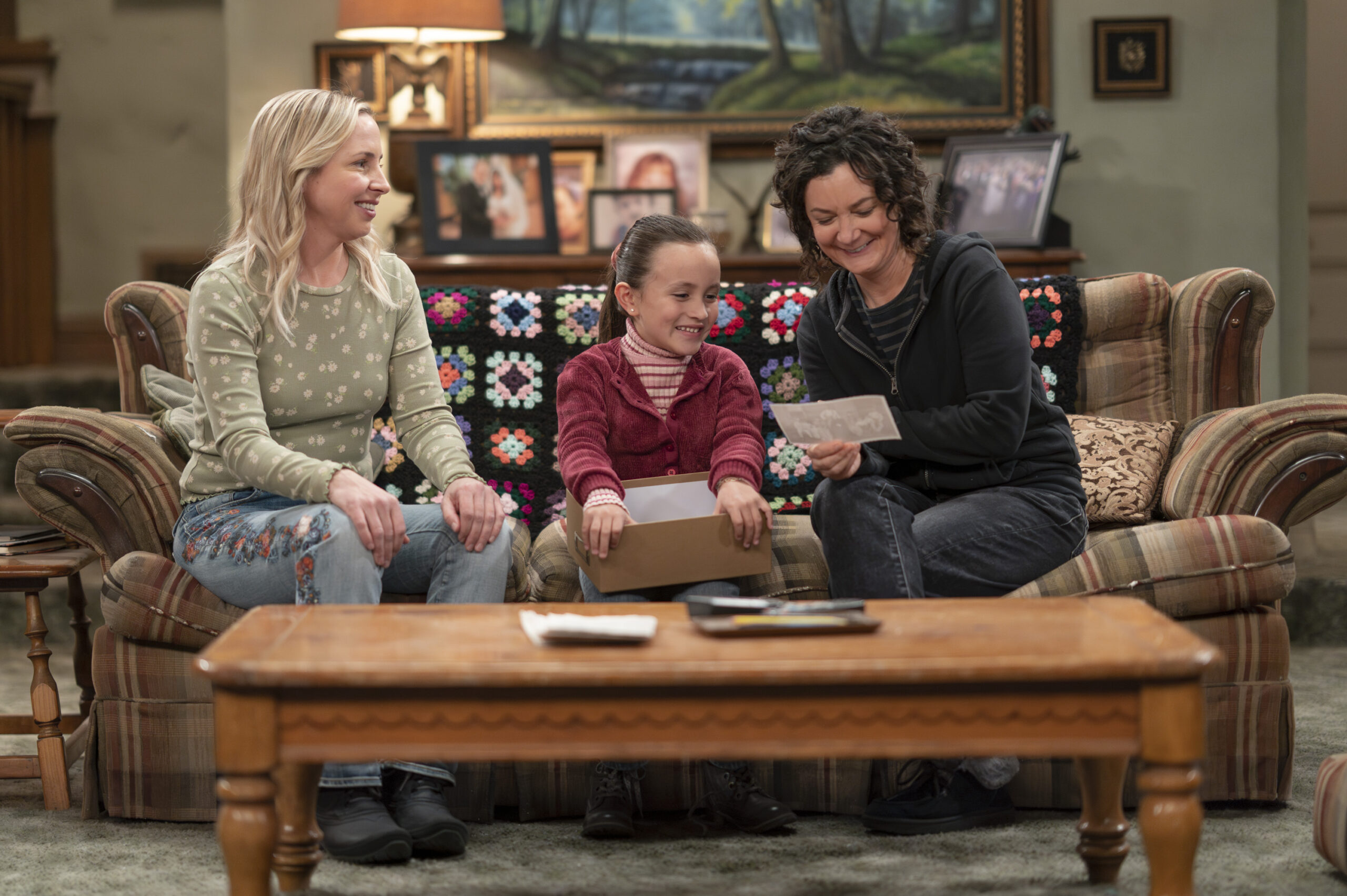 The Conners Season 6 Episode 7 - LECY GORANSON, CHARLOTTE SANCHEZ, SARA GILBERT sit on the Conner family couch looking at photos and smiling.