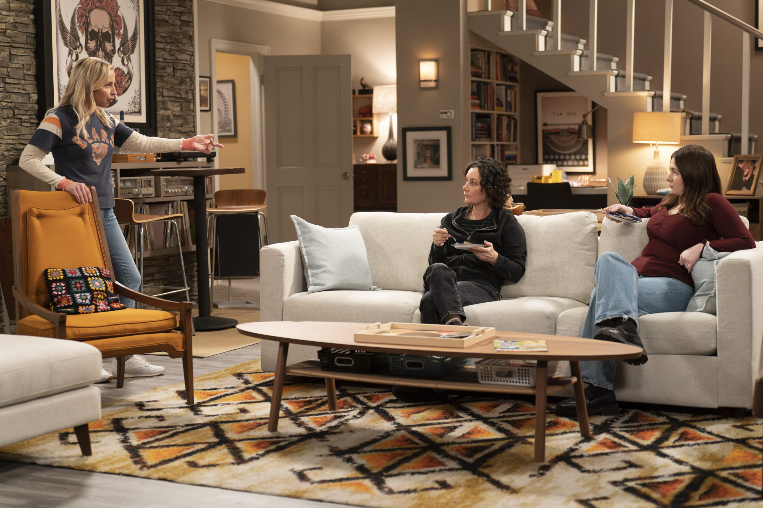 The Conners Season 6 Episode 7: LECY GORANSON and SARA GILBERT sit on their couch talking with EMMA KENNEY who stands nearby.