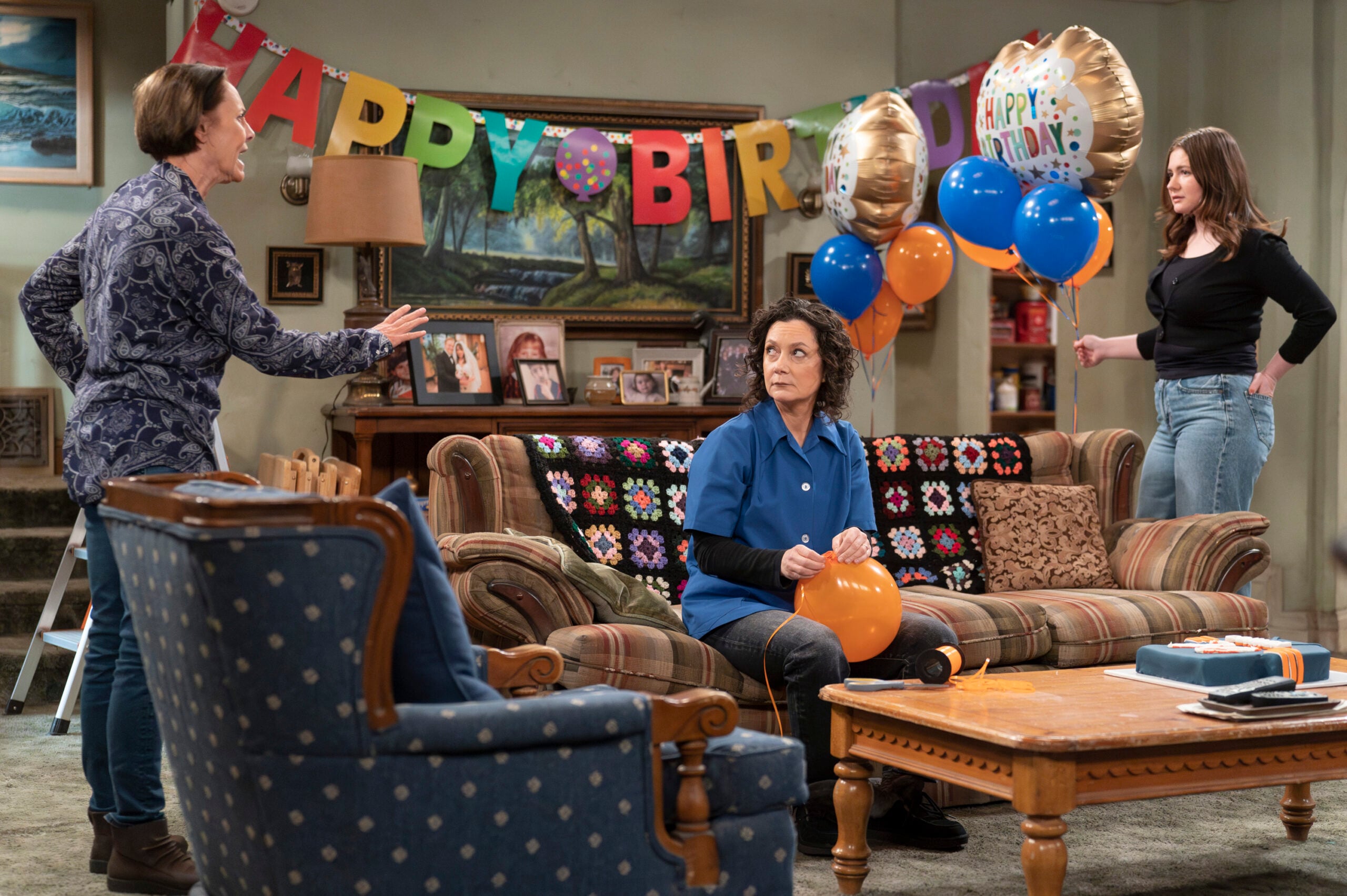 The Conners Season 6 Episode 8 - LAURIE METCALF, SARA GILBERT, EMMA KENNEY sit around the Conner living room with a Happy Birthday banner behind them.