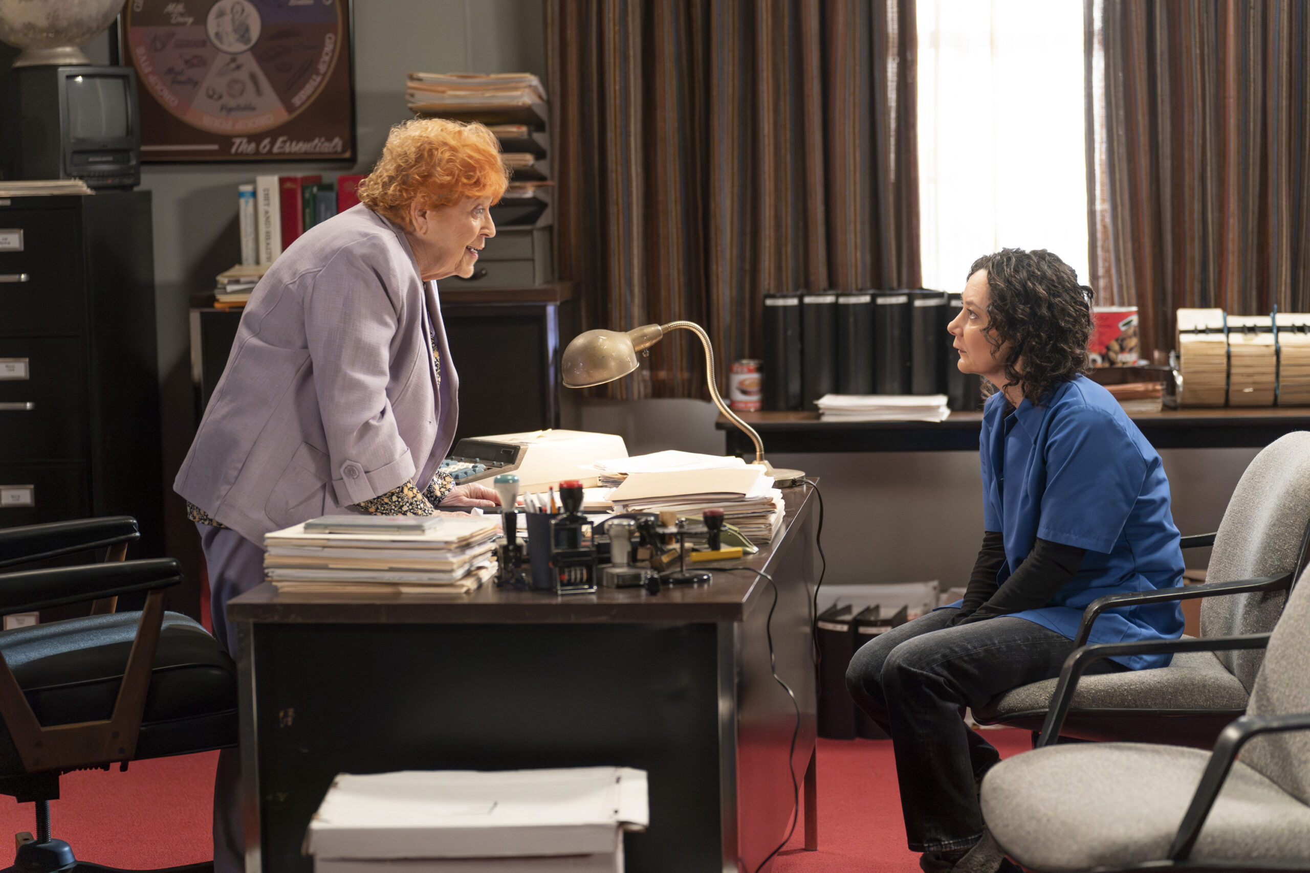 SUSAN BERGER, SARA GILBERT | Tell-Tale TV The Conners Season 6 Episode 8 - SUSAN BERGER stands behind her desk, being firm with SARA GILBERT who sits in a chair.