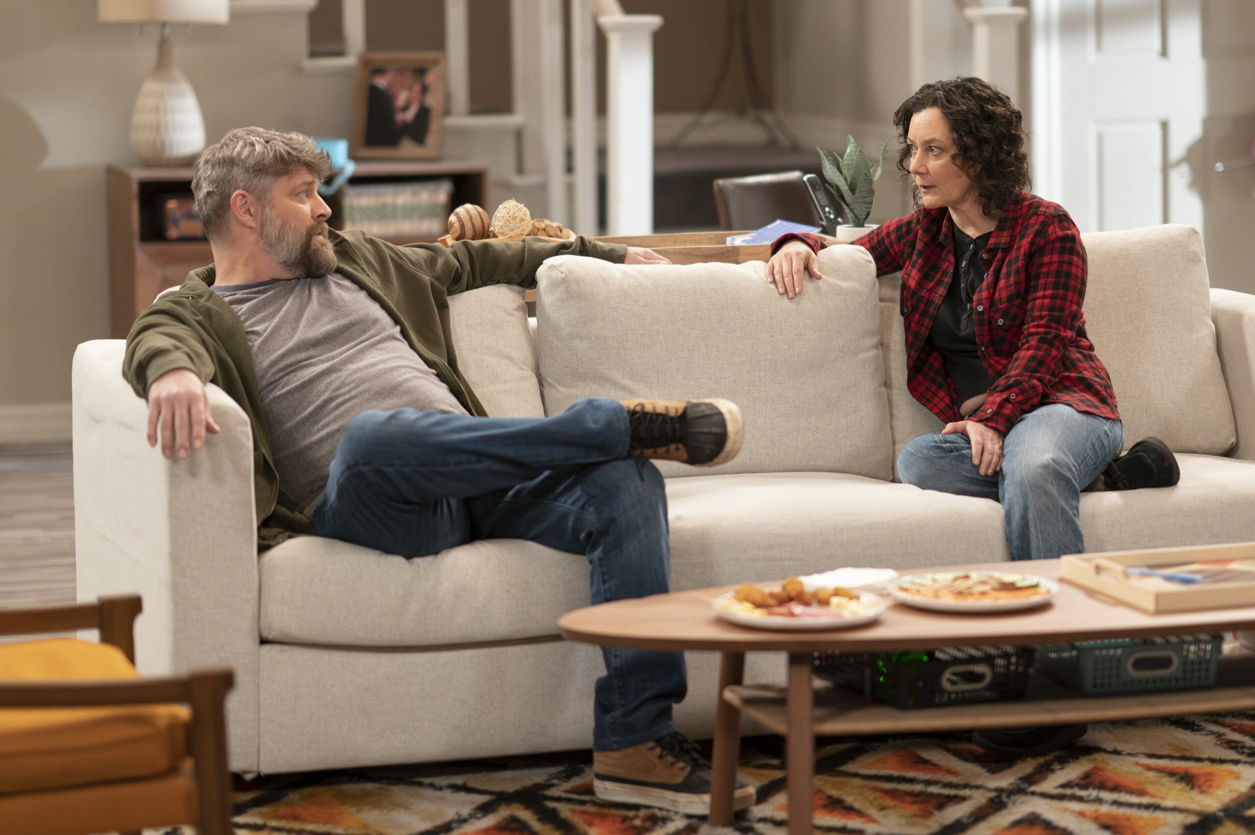 The Conners Season 6 Episode 8 - JAY R. FERGUSON and SARA GILBERT sit together on their couch and chat.