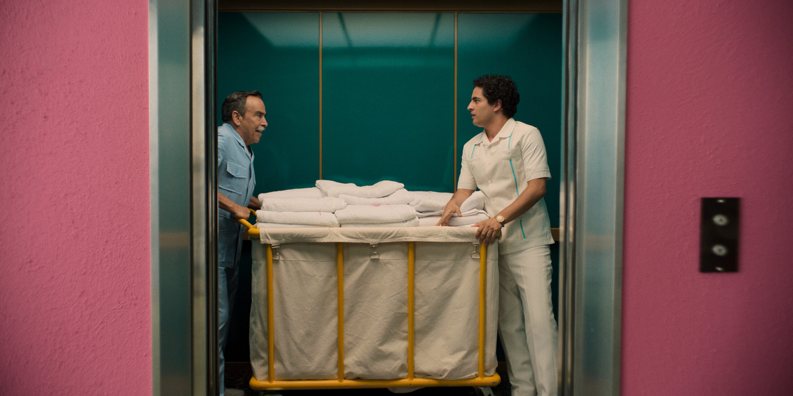 Still from Acapulco Season 3 Episode 3 of Damián Alcázar and Enrique Arrizon pictured from left to right standing in an elevator with a laundry cart in between them